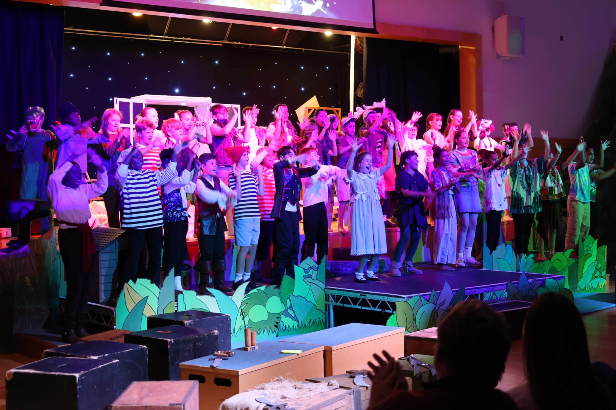 A large group of children in colourful costumes stand on stage performing a play, waving their hands. The stage is decorated with props and greenery, whilst an audience watches from the foreground.