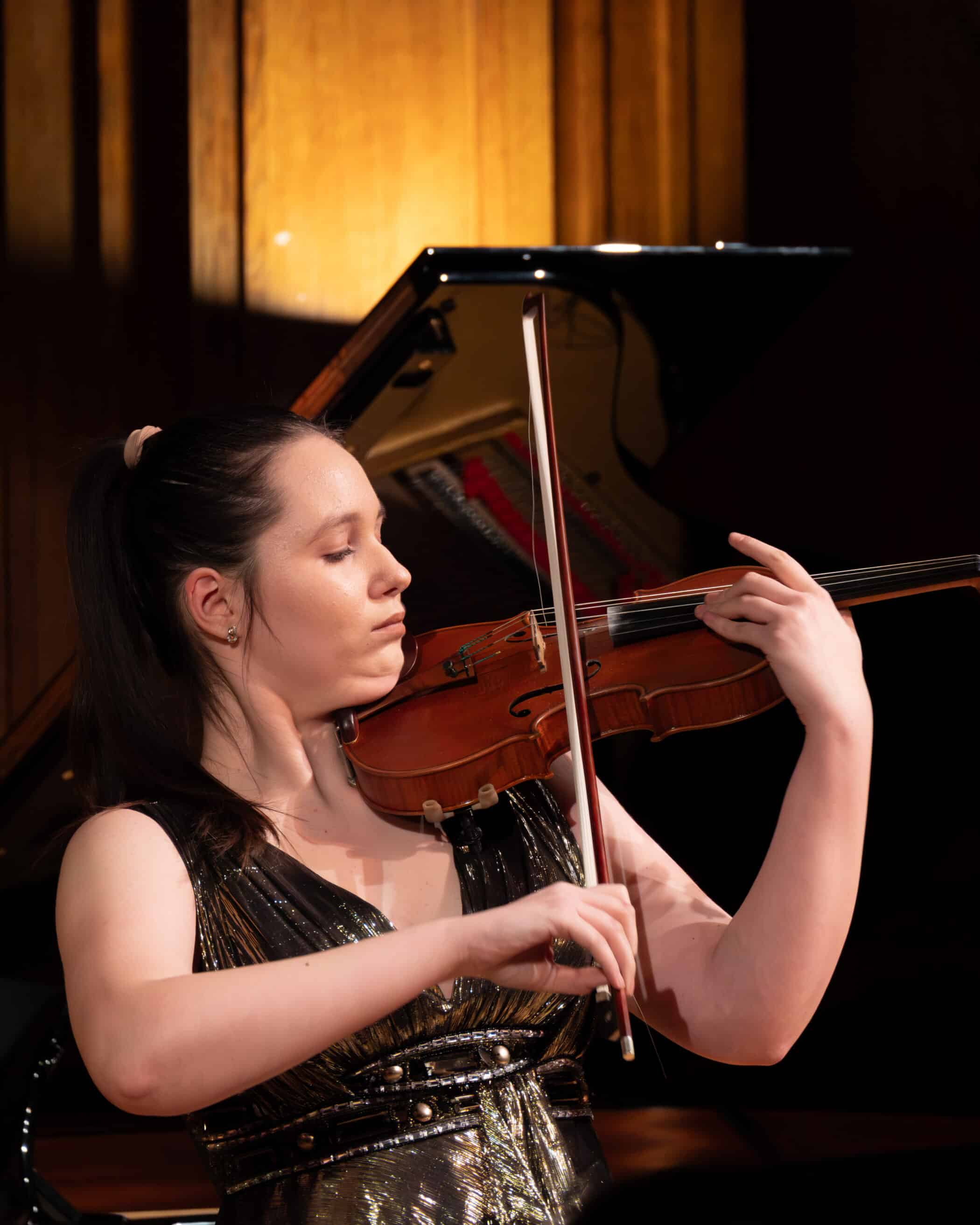 A woman in a sleeveless, metallic dress plays the violin with focus and emotion in front of a grand piano, under warm stage lighting.