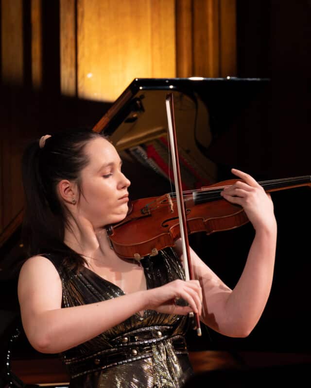 A woman in a sleeveless, metallic dress plays the violin with focus and emotion in front of a grand piano, under warm stage lighting.