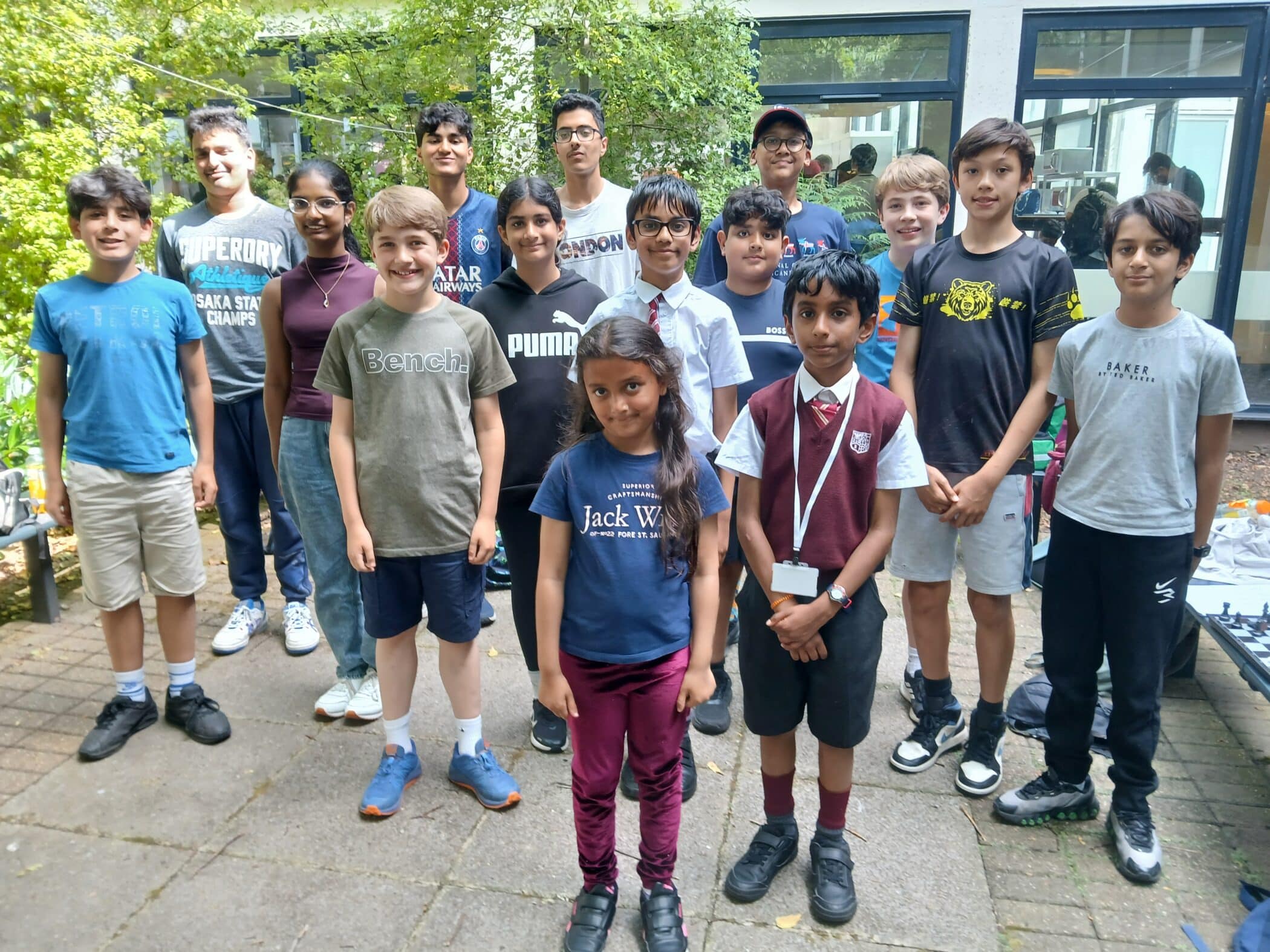 A group of sixteen children and teenagers stand together outside in a courtyard, posing and smiling for a group photo. Some wear casual clothes while a few are in school uniforms. Trees and a building are visible in the background.