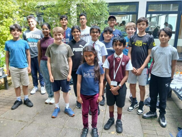 A group of sixteen children and teenagers stand together outside in a courtyard, posing and smiling for a group photo. Some wear casual clothes while a few are in school uniforms. Trees and a building are visible in the background.