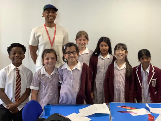 A group of seven children in school uniforms and a smiling adult wearing a white shirt and cap stand together in a classroom, with papers and stationery on a blue table in front of them.