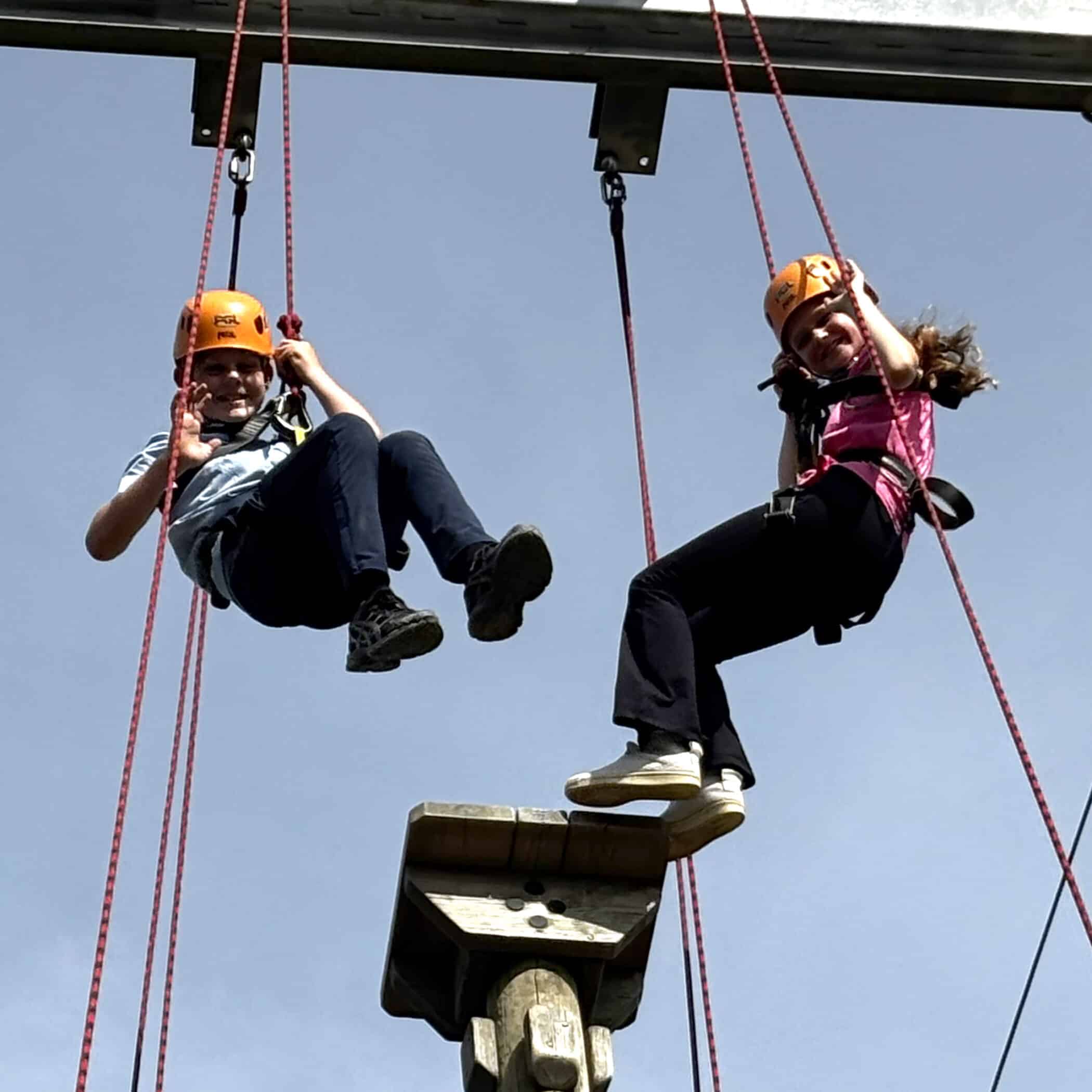 Two people wearing helmets and harnesses are suspended by ropes next to a tall wooden pole with foot platforms, participating in a high ropes course against a blue sky.