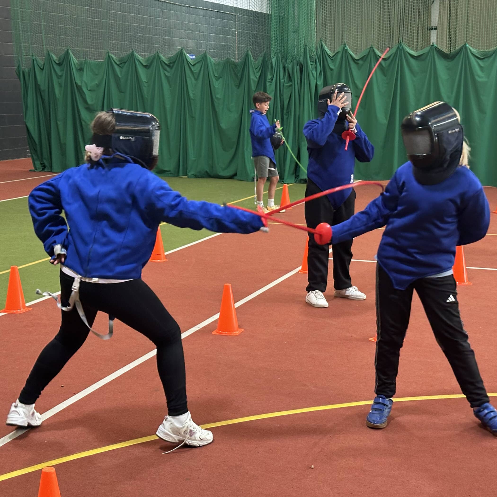 Two children in blue jackets and fencing masks spar with red swords in a gymnasium, surrounded by orange cones. Two others, also in safety gear, watch in the background. The floor is red with green curtains hanging behind.