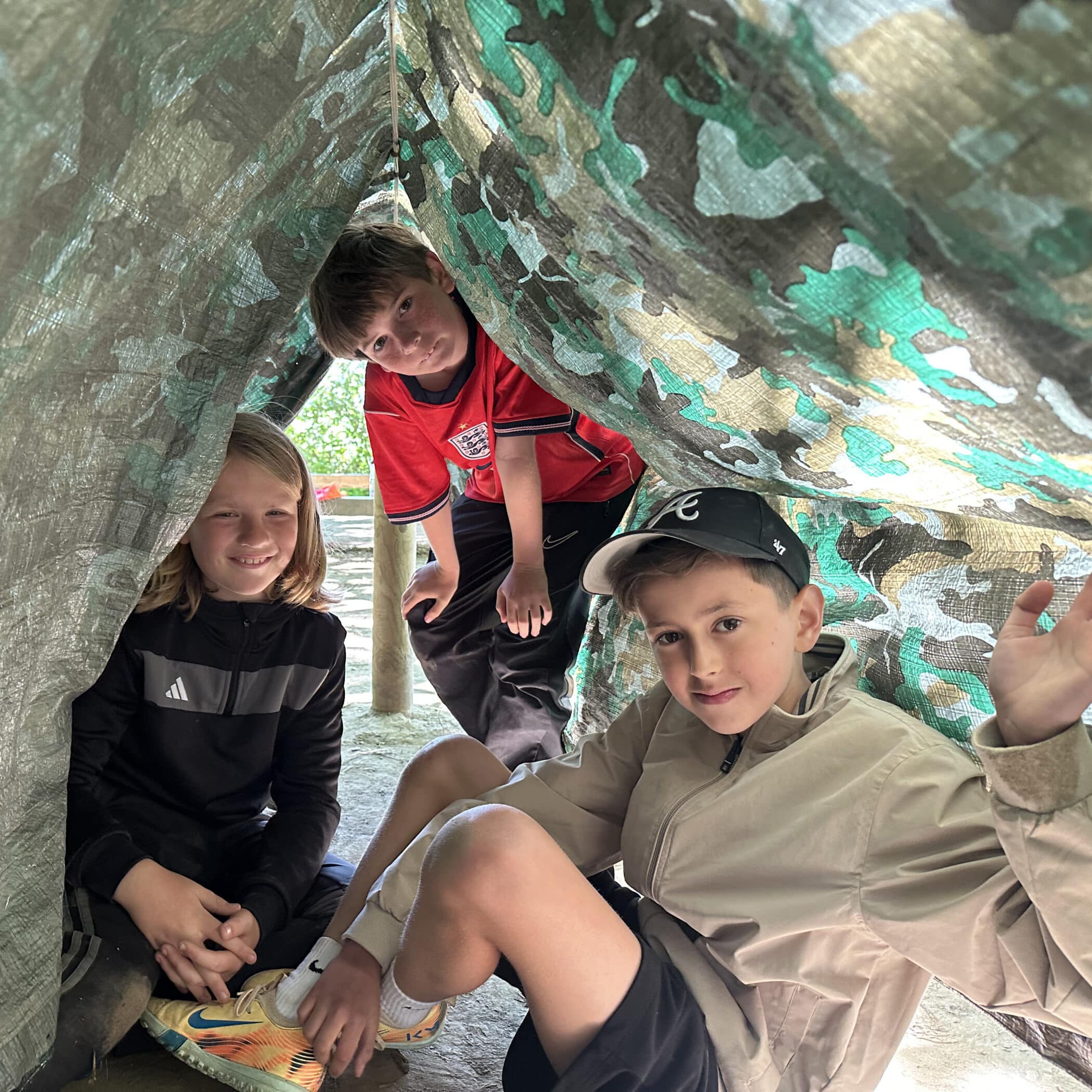 Three children sit and crouch together under a green and grey camouflage tarpaulin, smiling and looking at the camera. Daylight filters in, creating a cosy tent-like atmosphere.