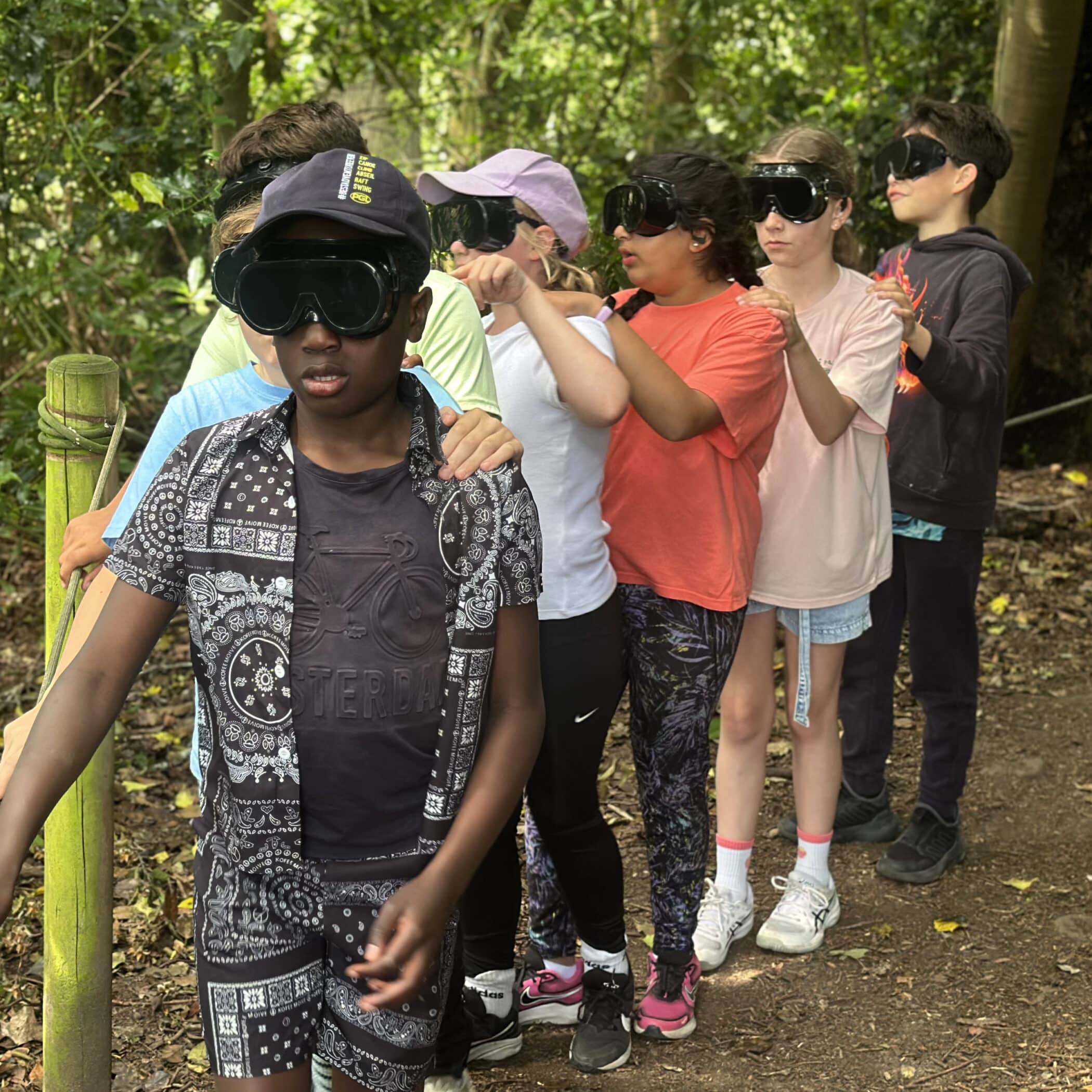 A group of children wearing dark goggles walk in a line through a wooded area, each holding onto the shoulders of the person in front of them, guided by a rope along a path.