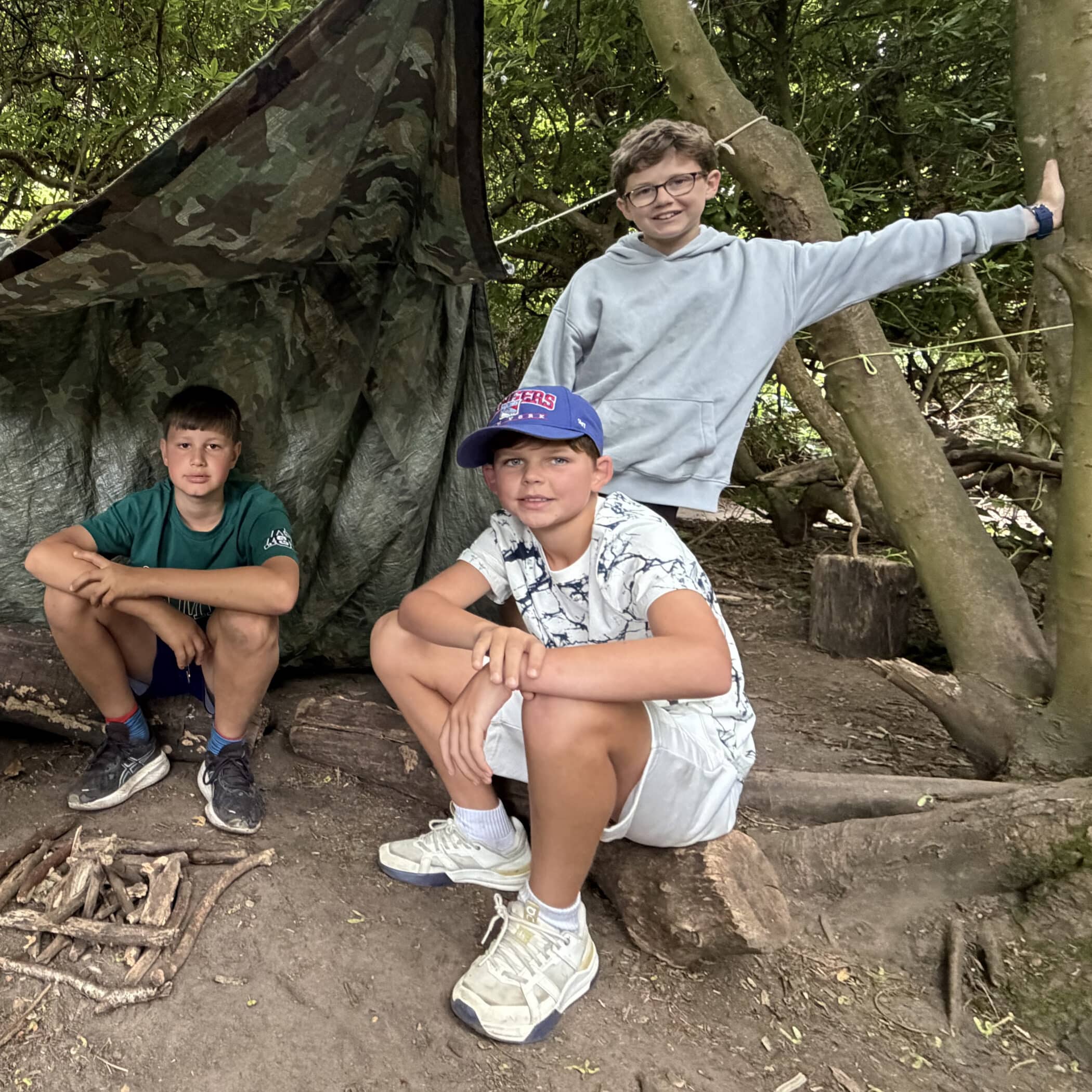 Three boys are outdoors in a wooded area, posing in front of a makeshift shelter made of camouflage tarpaulin and sticks. Two boys sit on logs whilst one stands, all smiling at the camera.
