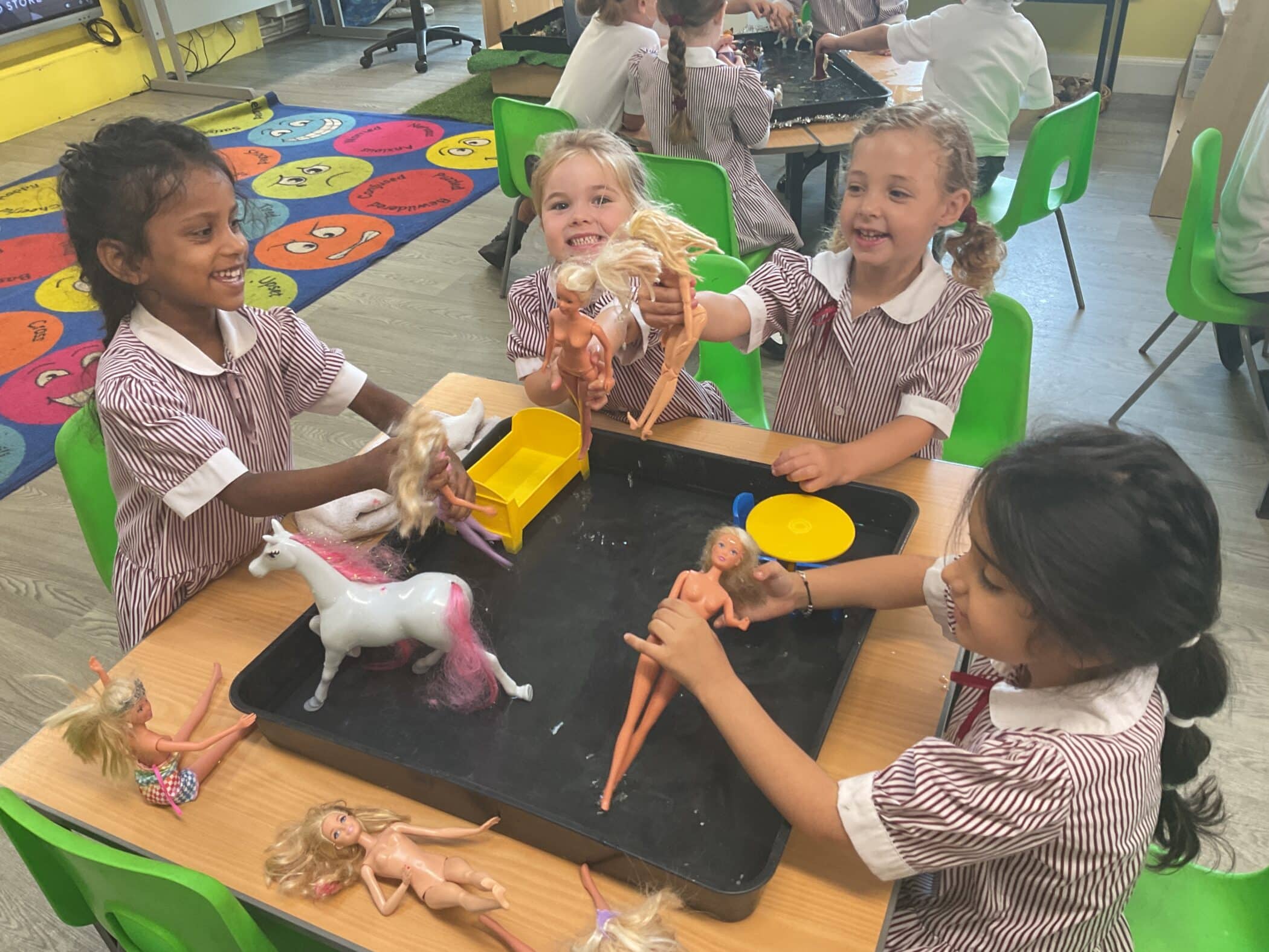 Four young girls in striped school uniforms sit around a table, smiling and playing with dolls and a toy unicorn. They appear to be enjoying an indoor classroom activity together.