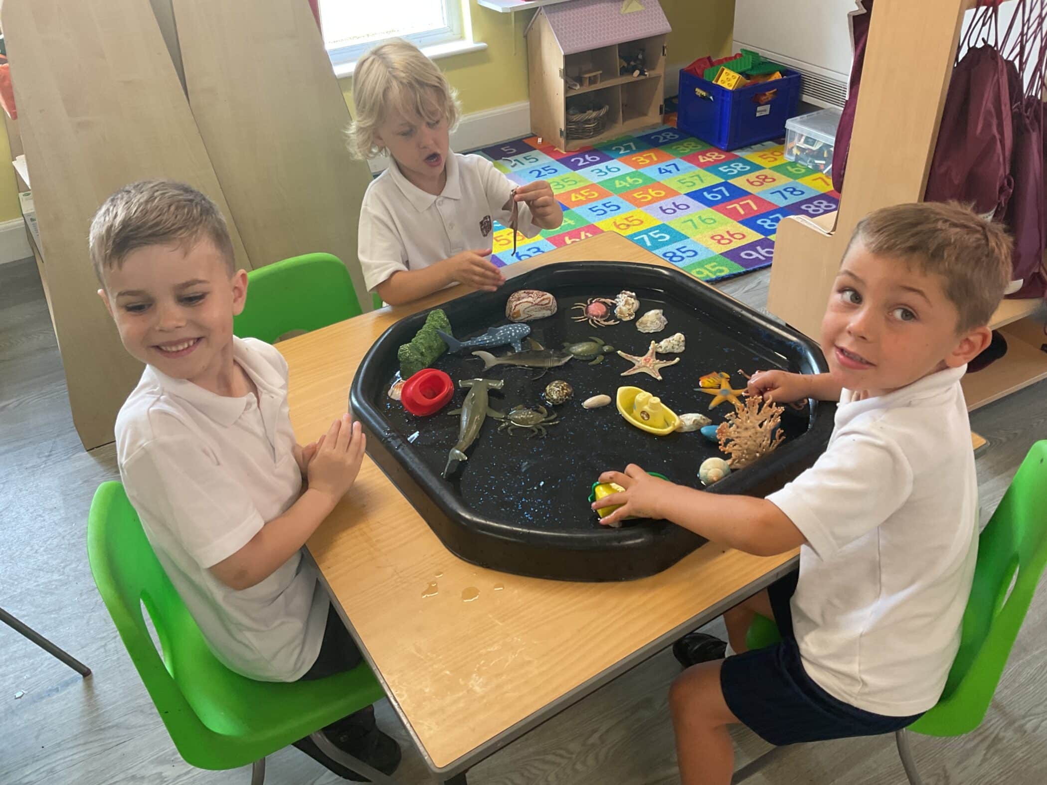 Three young boys in white shirts sit around a sensory play table with sea-themed toys and shells in a colourful classroom. Two boys are smiling at the camera while one is playing with a toy.