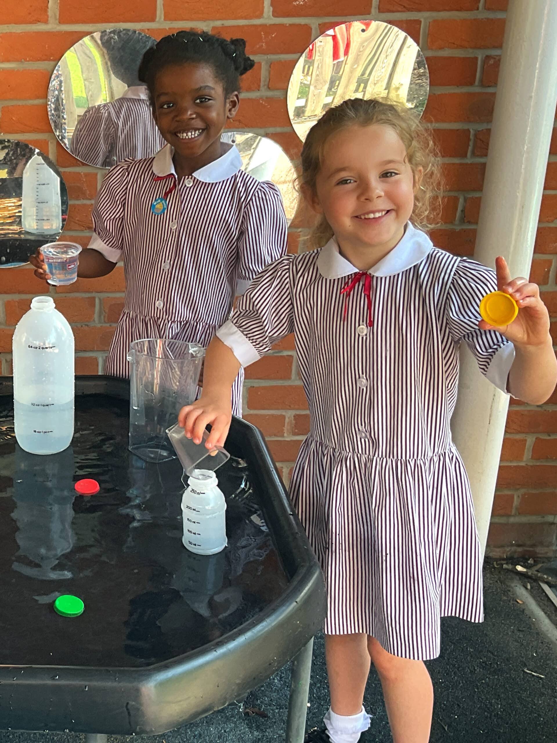 Two young girls in striped uniforms smile whilst playing with water and containers at a sensory table. They are outdoors, standing in front of a brick wall with round mirrors attached to it.