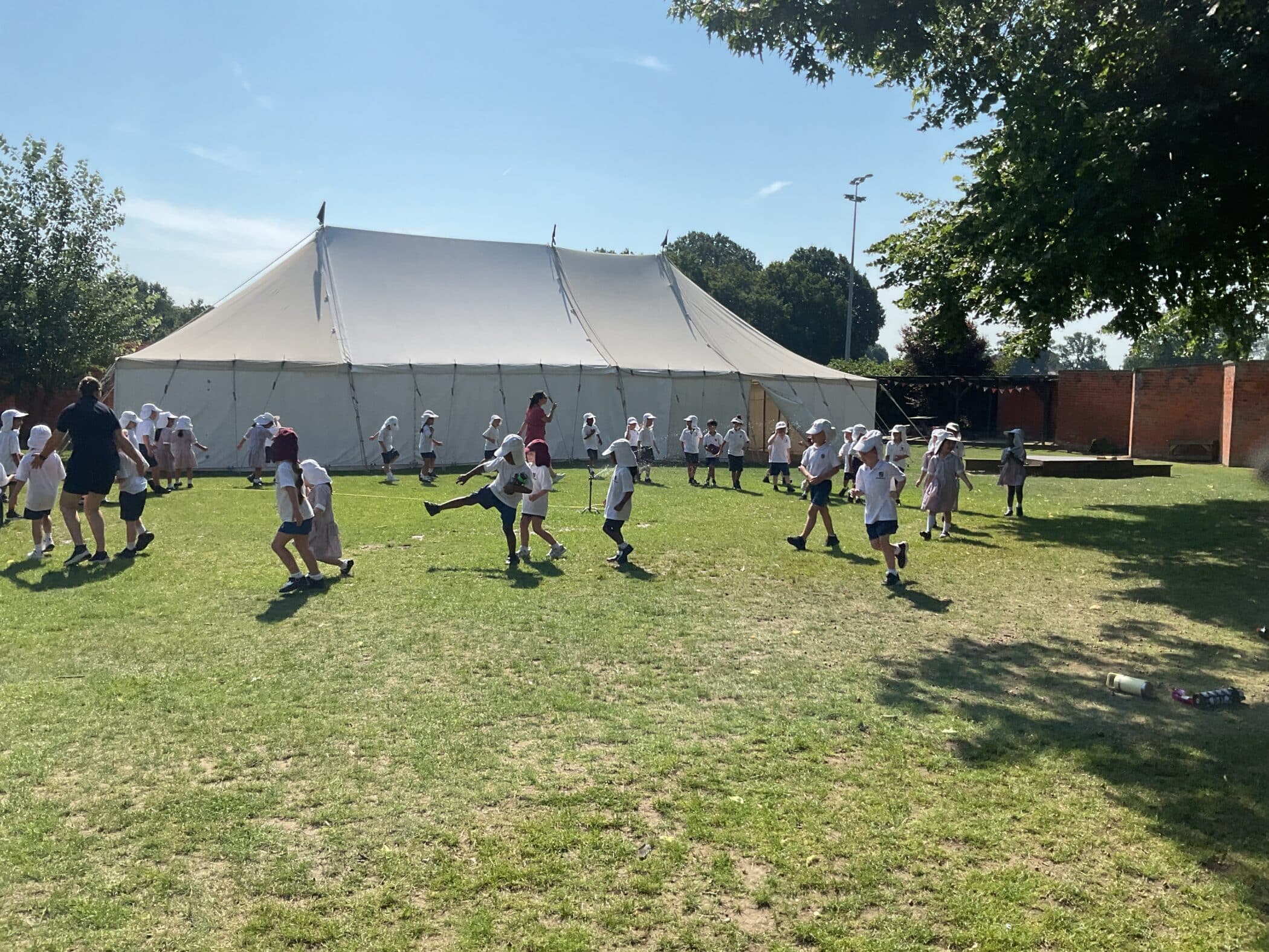 Children in white uniforms and hats play on a sunny grassy field near a large white marquee. Two adults supervise, and trees provide some shade. The sky is clear and blue.
