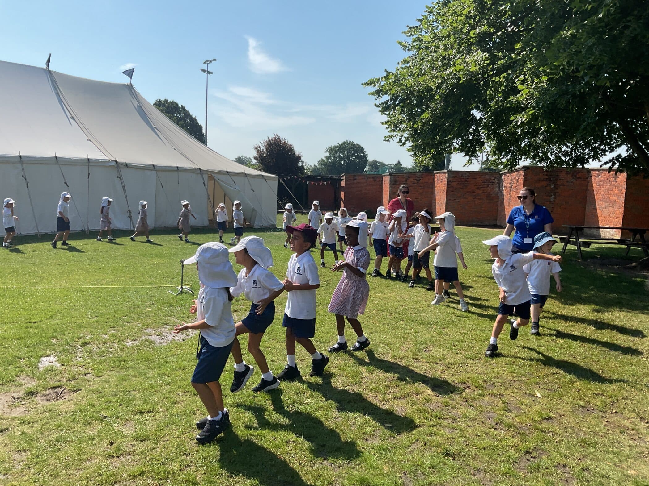 A group of young children wearing sun hats and light clothing play outside on grass near a large white marquee, supervised by an adult under a sunny sky with some shade from nearby trees.