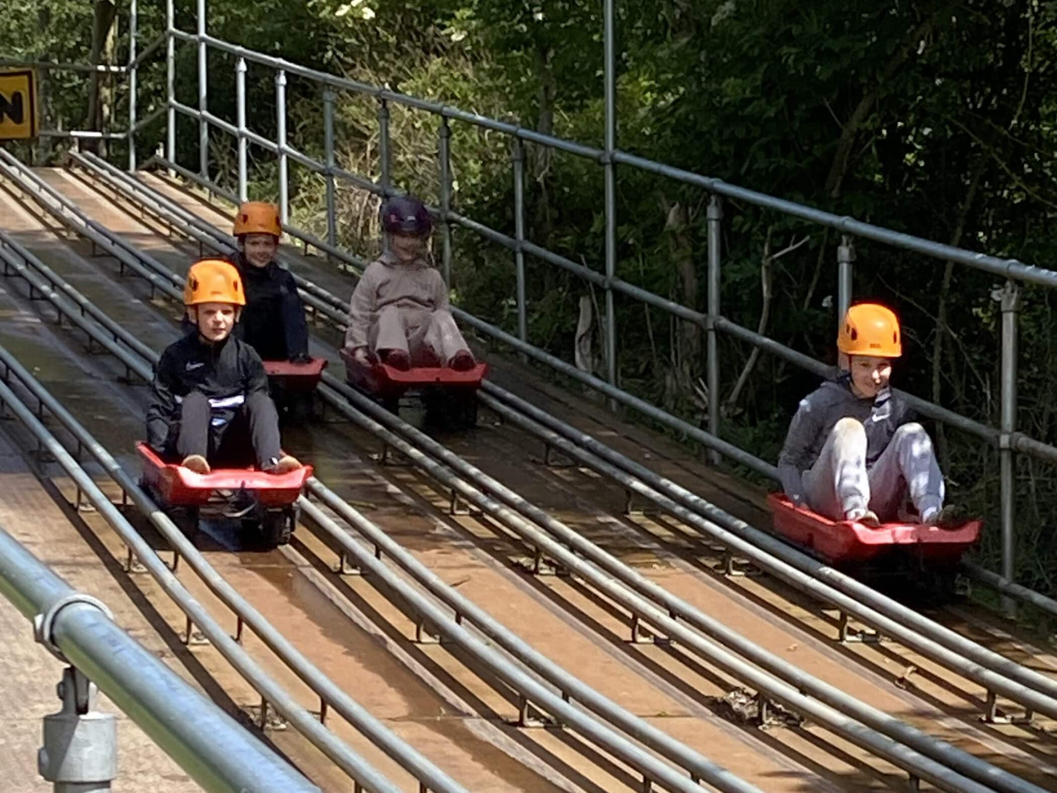 Four children wearing helmets ride individual red carts down parallel metal tracks on an outdoor luge course, surrounded by greenery and trees. They appear focused and are dressed in casual clothing.