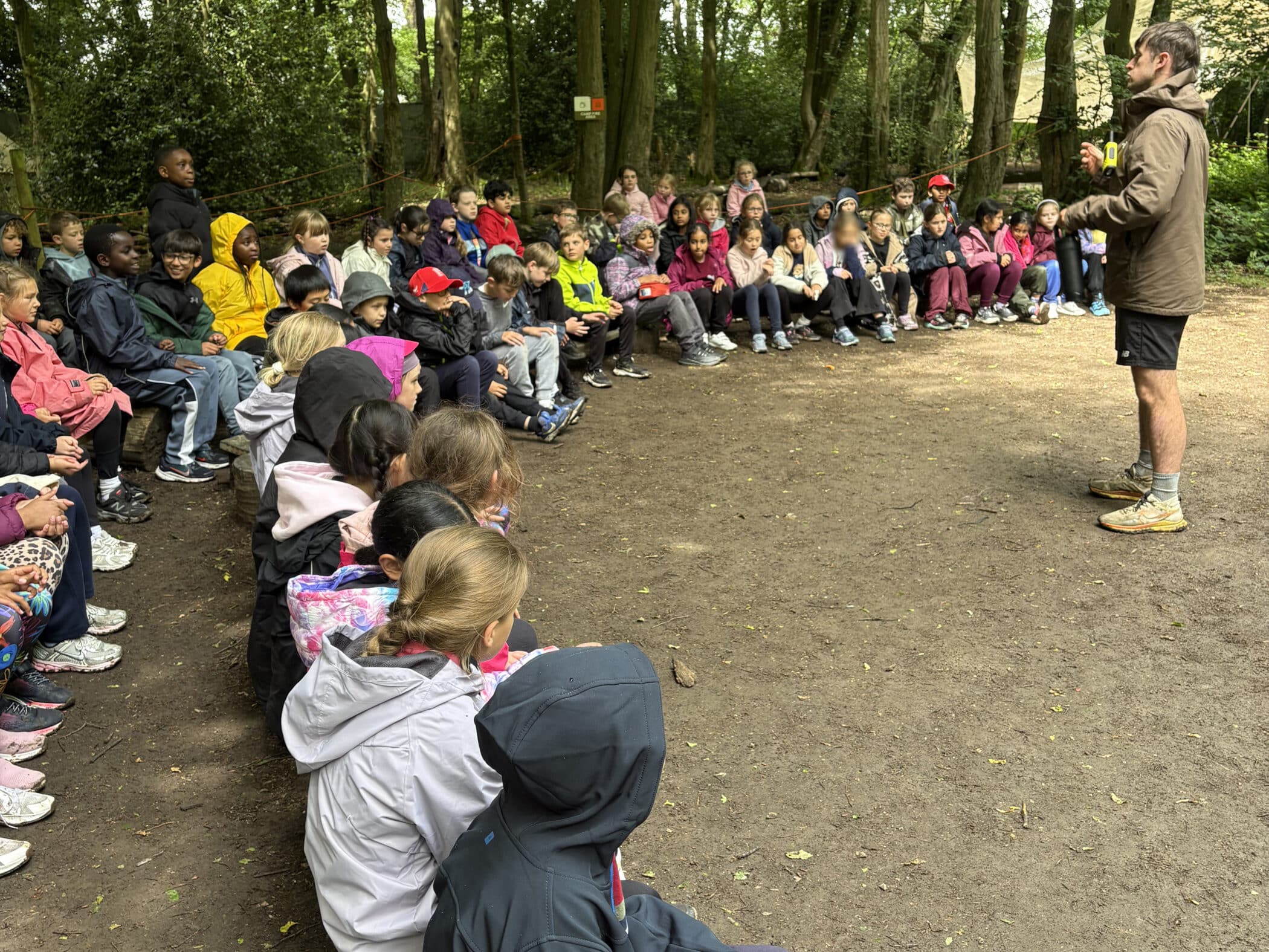 A group of children in outdoor clothing sit on benches in a wooded area, listening attentively to an adult standing in front of them and speaking. Trees and greenery surround the gathering.
