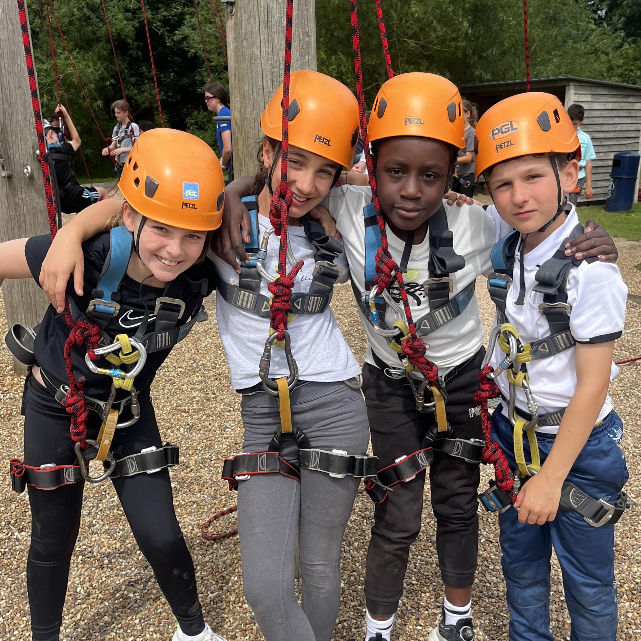 Four children wearing orange helmets and harnesses stand close together outdoors, smiling at the camera. They appear ready for a climbing or ropes course activity, with ropes and wooden structures in the background.