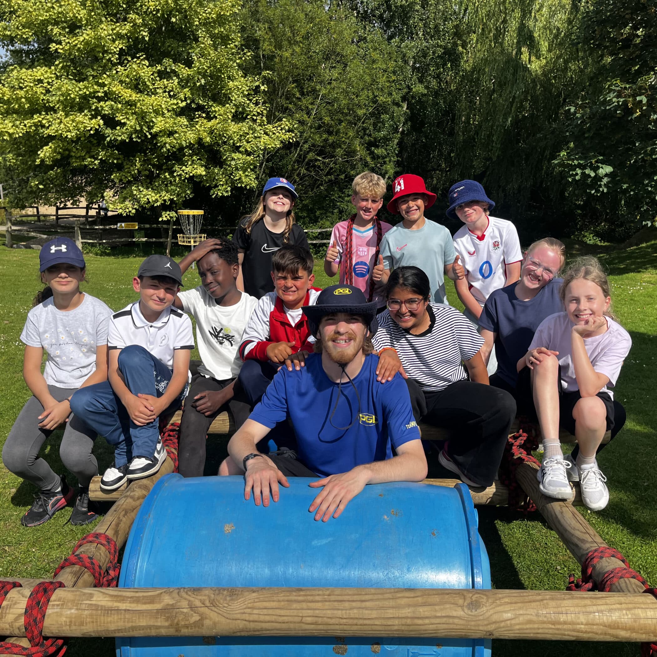 A group of children and an adult sit and stand around a blue barrel outdoors on a sunny day, smiling at the camera. Trees and grass surround them, and everyone looks happy and relaxed.