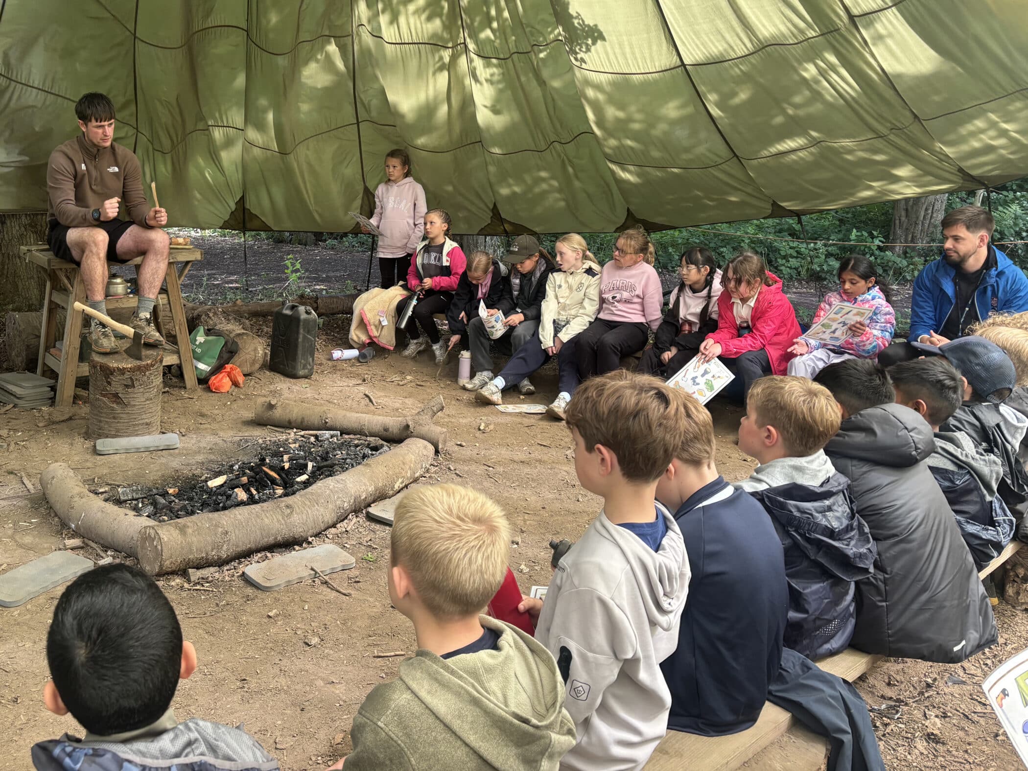 A group of children sit on benches under a large green canopy, listening to a man demonstrating survival skills near a stone-lined outdoor fire pit in a wooded area. Another adult sits among the children.
