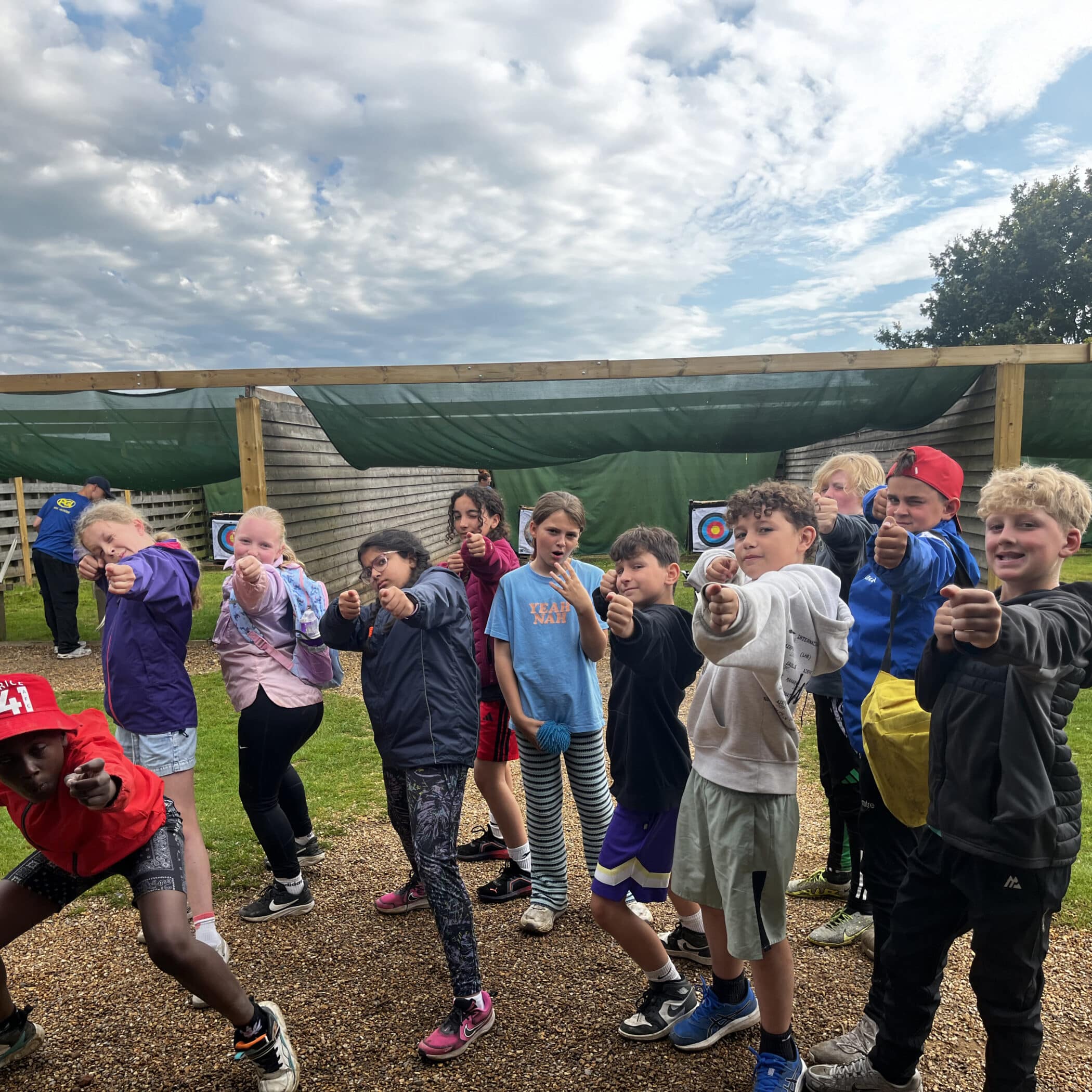 A group of children pose outdoors at an archery range, pointing their fingers playfully like toy guns towards the camera. They stand on gravel with targets visible in the background under a cloudy sky.