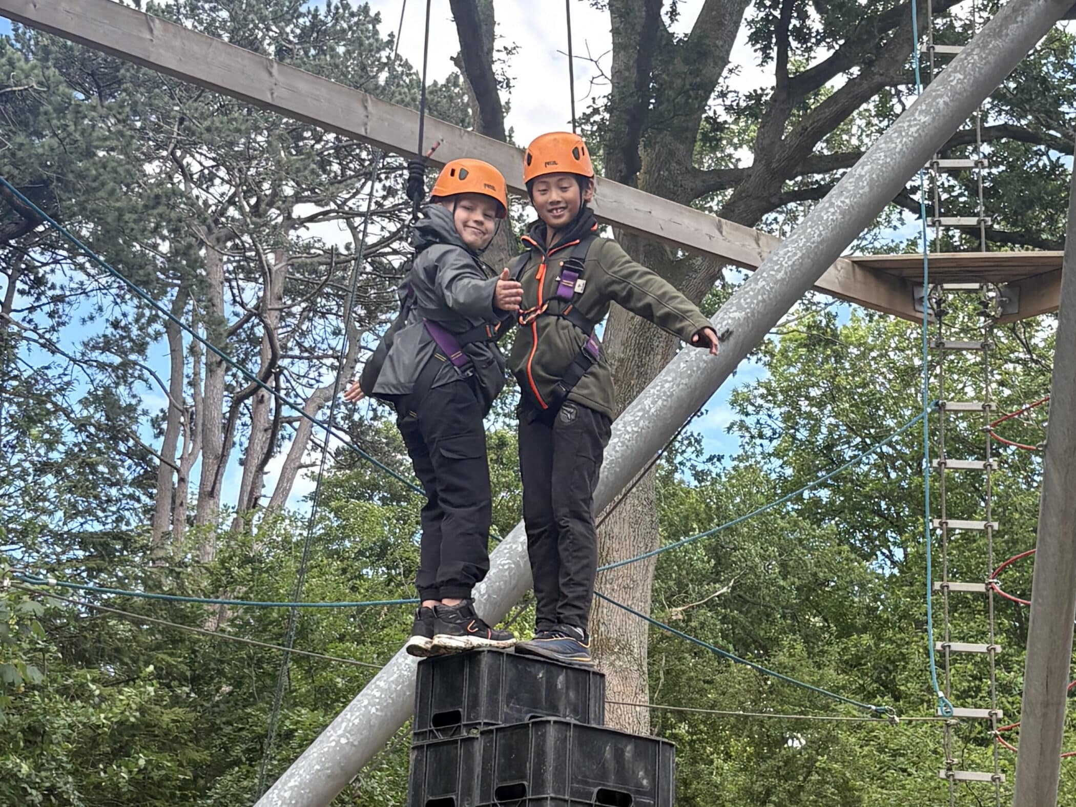 Two children wearing helmets and harnesses stand on stacked crates during an outdoor climbing activity, giving a thumbs-up and smiling, with trees and ropes in the background.