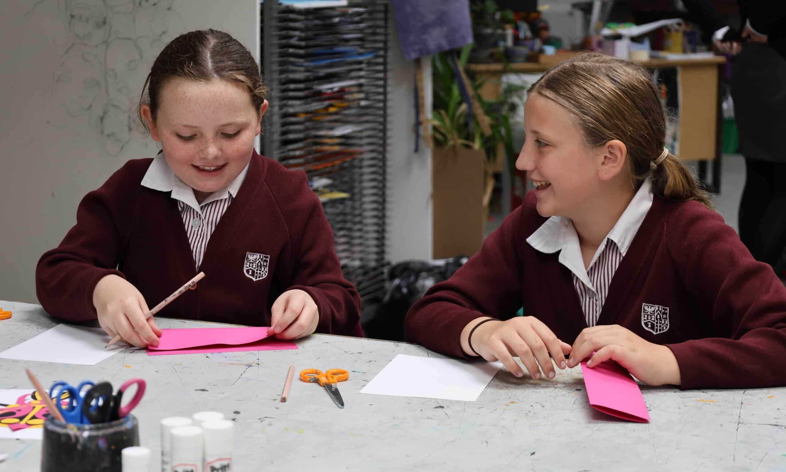 Two girls in maroon school uniforms sit at a table, smiling and working on a craft project—part of a Prep School to Senior School Transition activity—with pink paper, pencils, scissors, glue sticks, and other supplies spread out in the classroom.