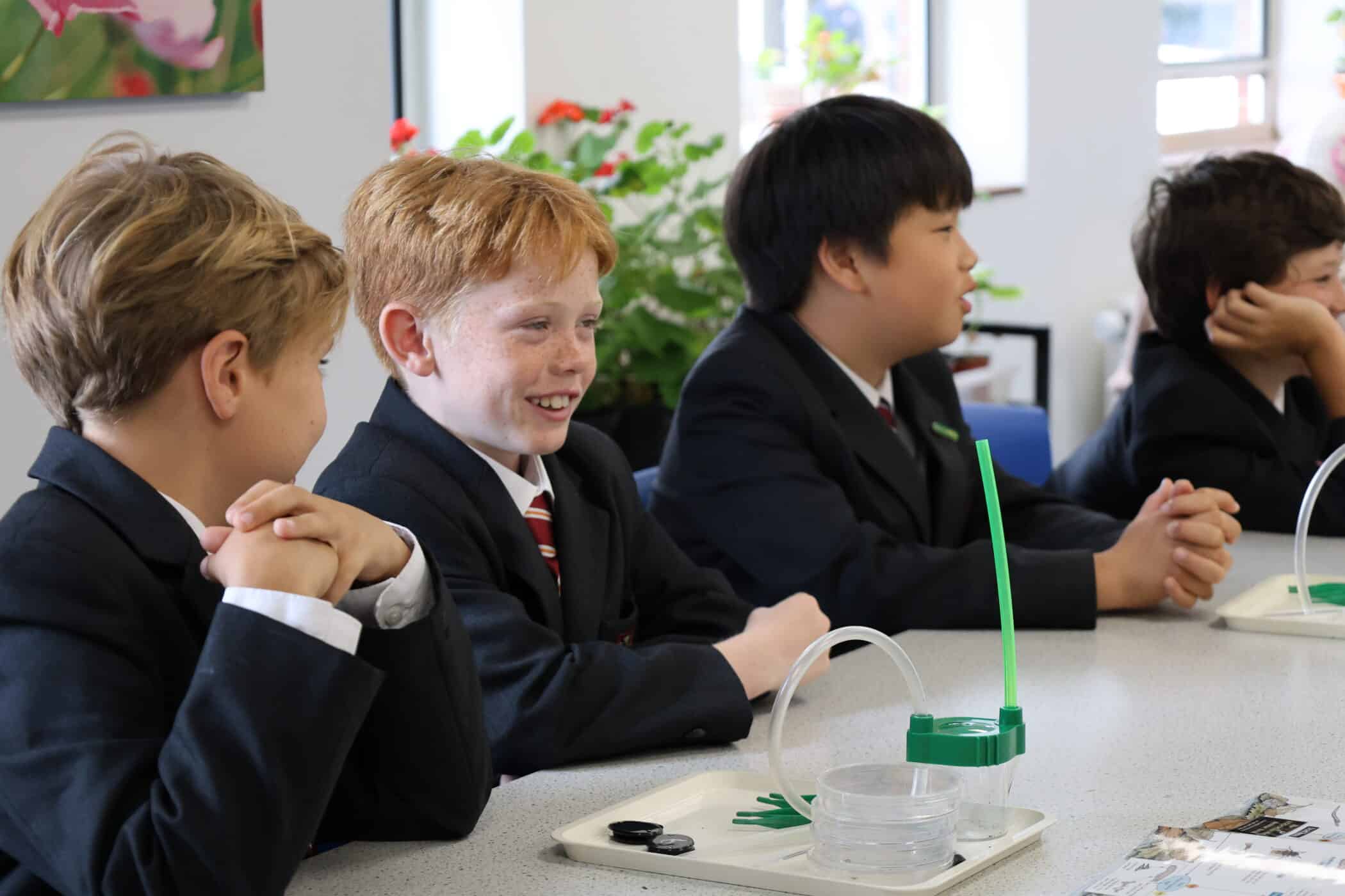 Four schoolboys in uniforms sit at a table with science equipment, including petri dishes and tubing, engaged in a classroom activity—an inspiring glimpse of Prep School to Senior School Transition. One boy smiles, with plants visible in the background.