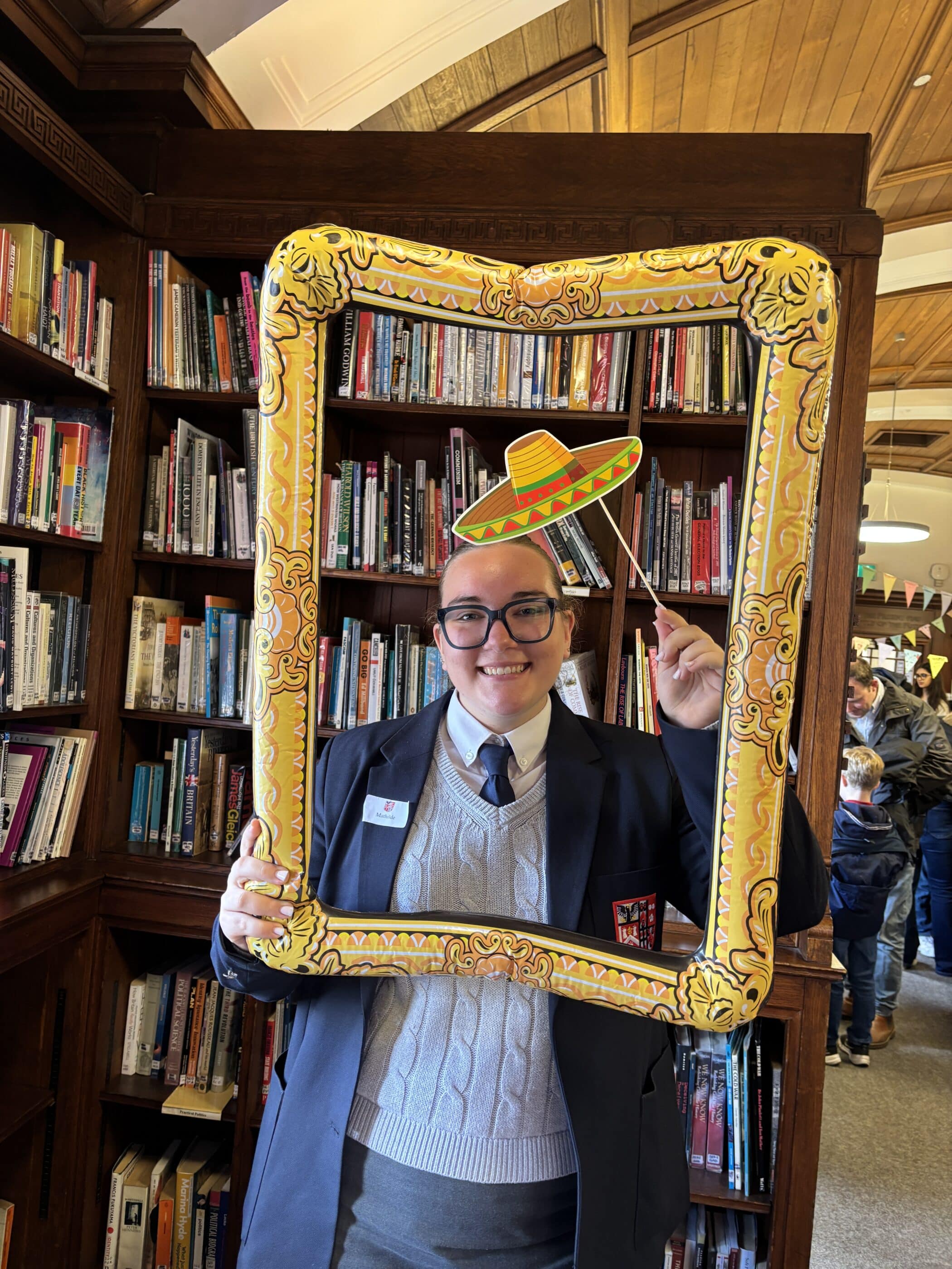A person in a school uniform smiles whilst holding a decorative yellow picture frame and a paper sombrero on a stick during Open Events in front of bookshelves at a library or bookshop. Other people are visible in the background.