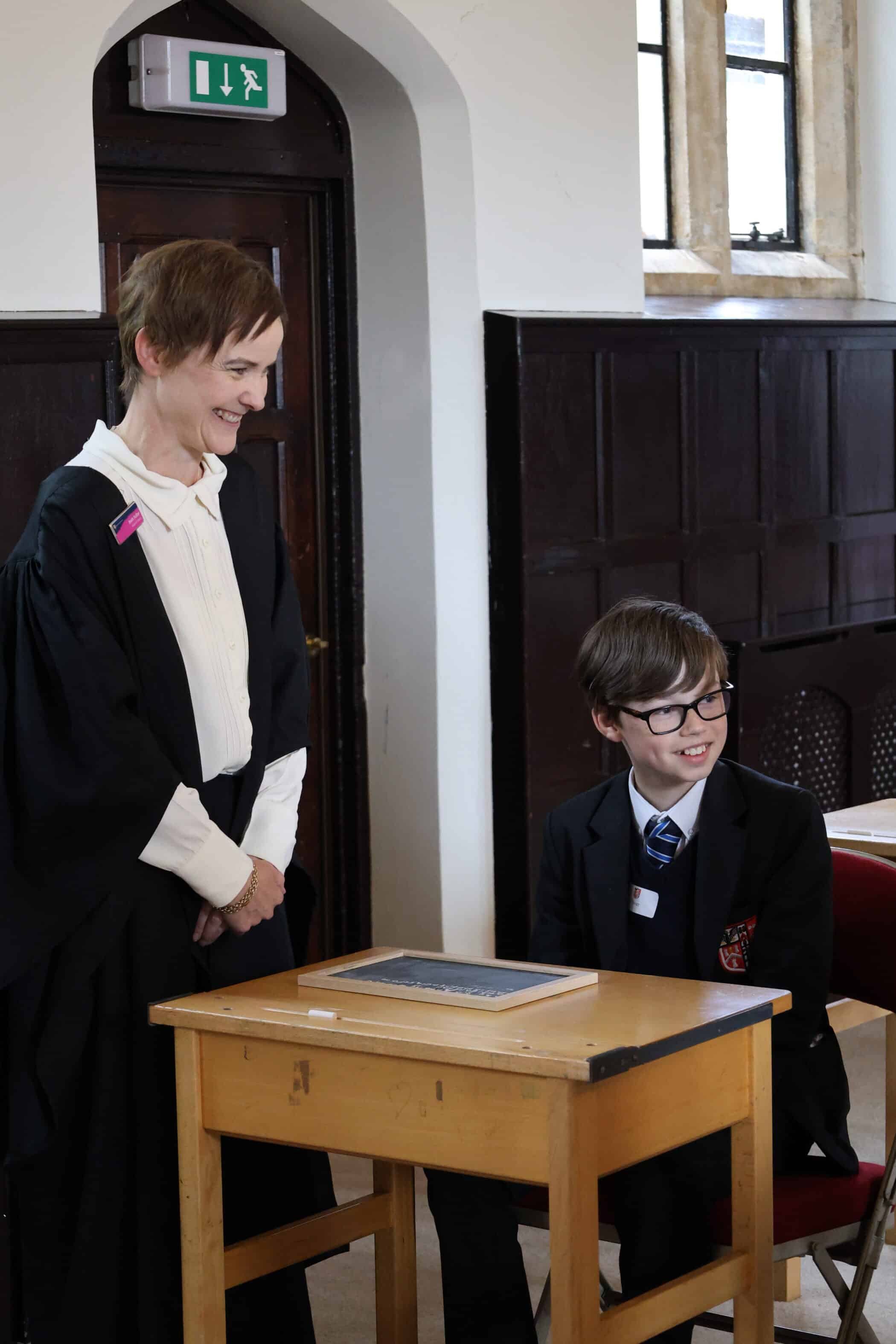 A teacher in academic robes stands smiling beside a seated pupil in a school uniform at a wooden desk, in a classroom with dark wood panelling and large windows—capturing the welcoming spirit of our Open Events.