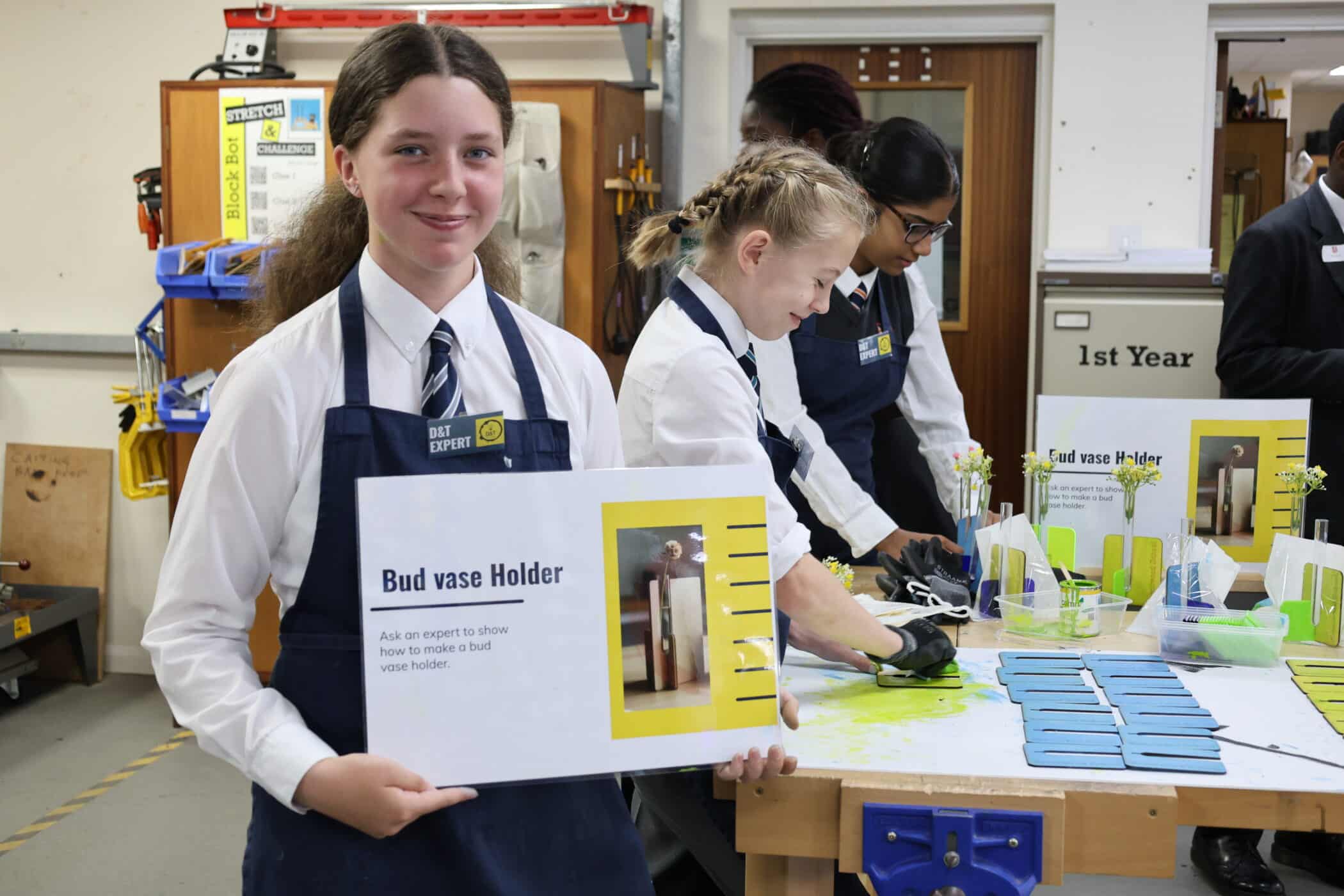 A student in a school workshop smiles and holds a sign reading Bud vase Holder during Open Events. Other students in uniforms and aprons work on craft projects at tables in the background.