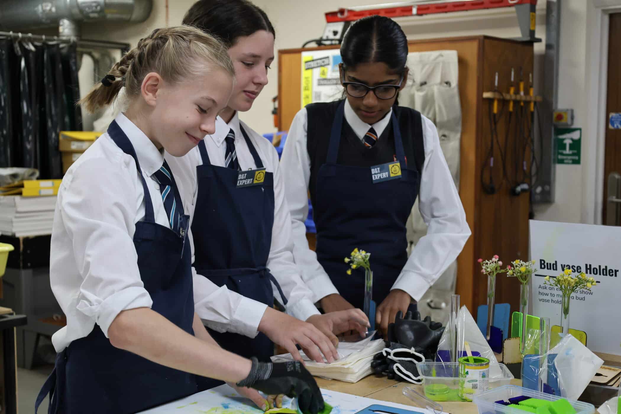 Three students in school uniforms and aprons work on a creative project at a table during one of the Open Events, smiling and engaged. A sign reads Bud Vase Holder, with examples and tools visible around them.