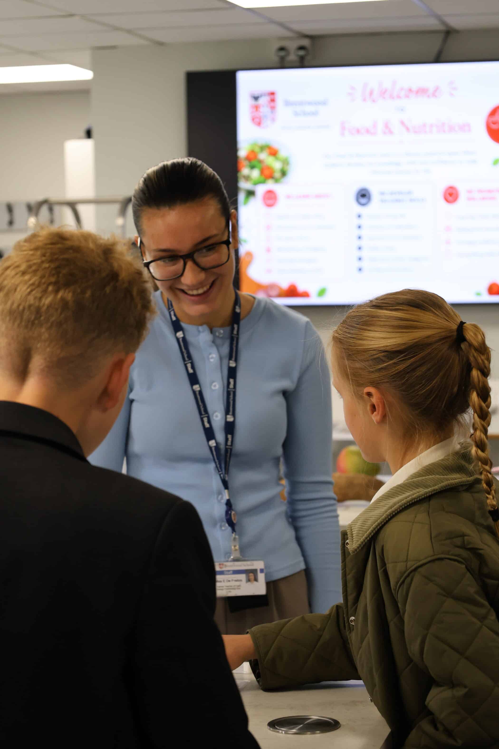A woman wearing glasses and a blue top smiles whilst talking to two children in a classroom. A welcome sign for Food & Nutrition is visible on a screen in the background, highlighting Open Events for families.