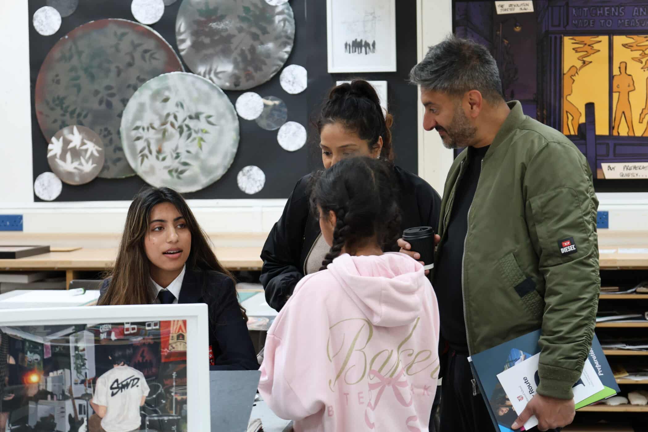 A student in uniform sits at a desk speaking to three adults, one holding a coffee cup and books, during Open Events in a classroom decorated with circular art pieces and drawings on the wall.