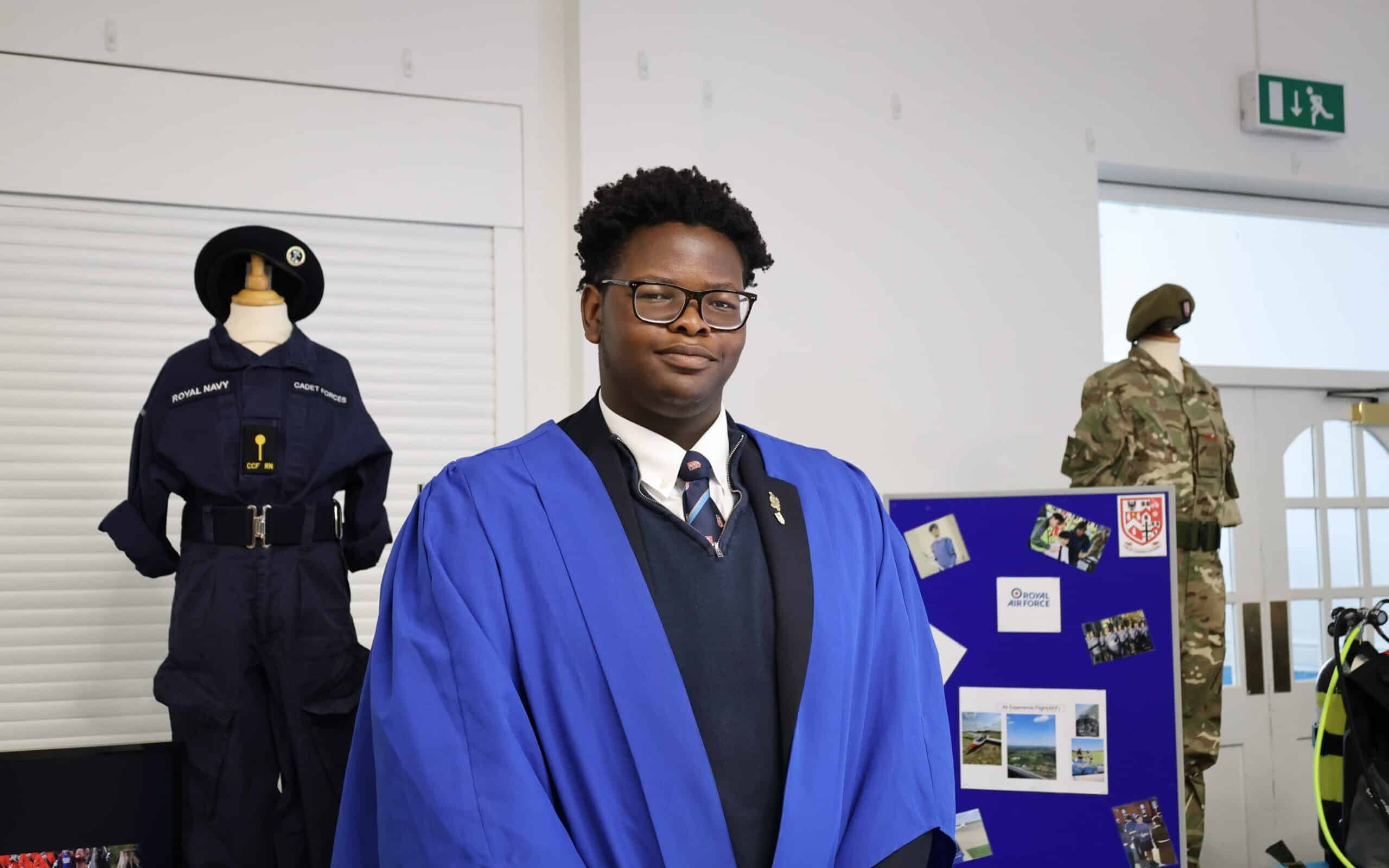 A student in a blue graduation gown stands indoors, with military uniforms displayed on mannequins and a blue information board detailing sixth form stay or move school options in the background.