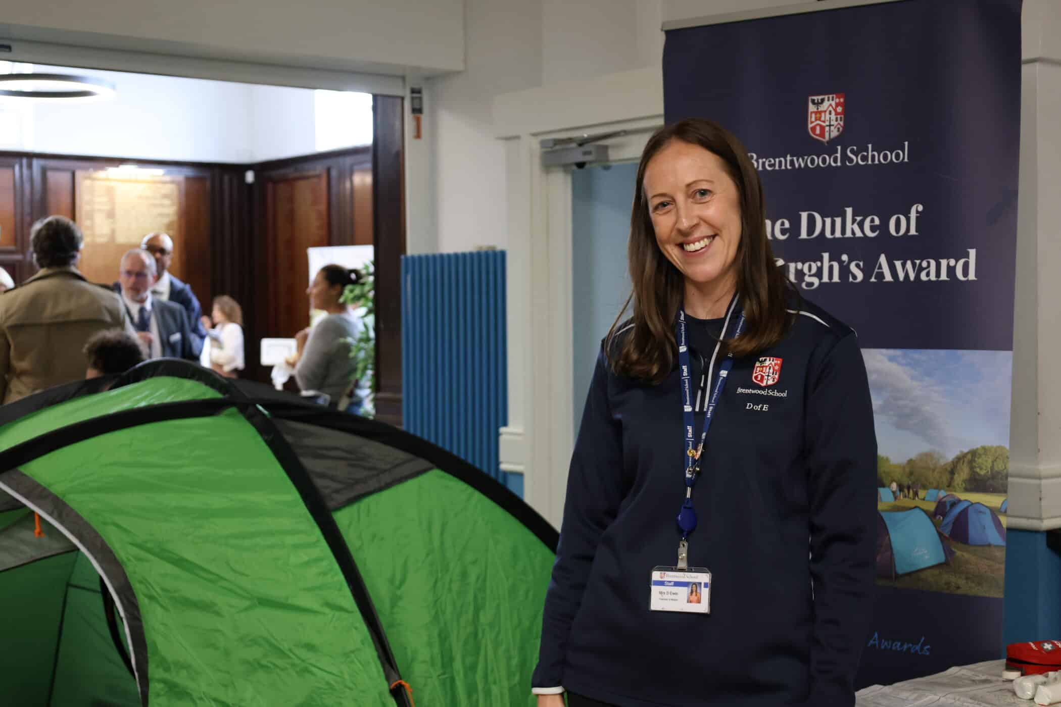 A woman smiles beside a green tent at an indoor Open Events gathering. She wears a navy sweatshirt and lanyard, standing before Brentwood School and The Duke of Edinburgh’s Award banners, as people mingle in the background.