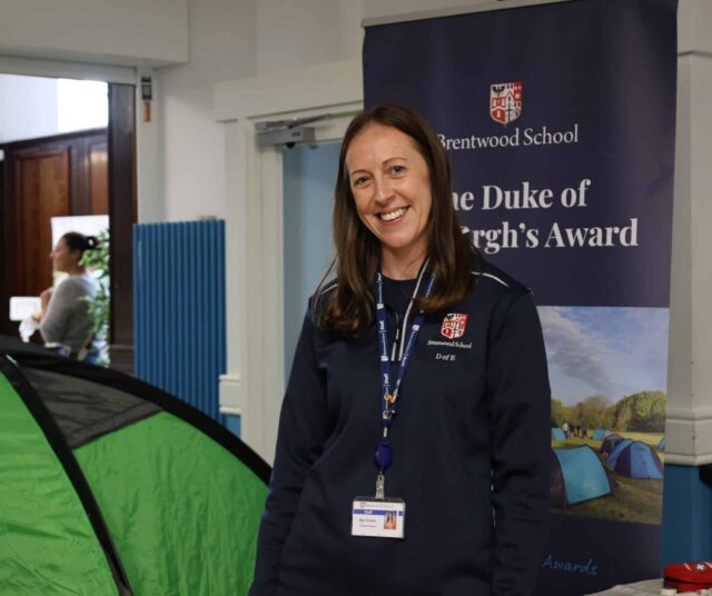 A woman smiles beside a green tent at an indoor Open Events gathering. The member of staff wears a navy sweatshirt and lanyard, standing before Brentwood School and The Duke of Edinburgh’s Award banners, as people mingle in the background.