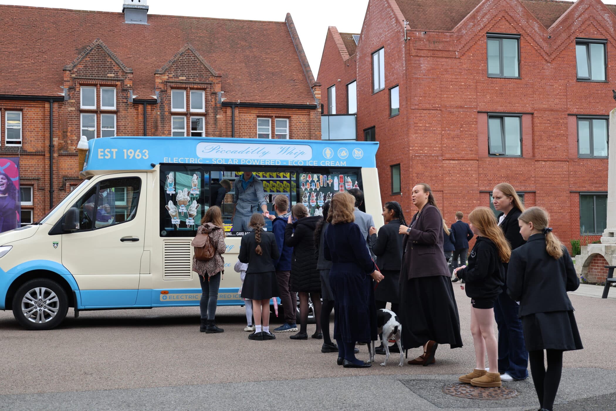 A group of people, mostly children in school uniforms, gather around a blue and white ice cream van parked on a street in front of red brick buildings, enjoying an open event. An adult with a dog stands nearby.