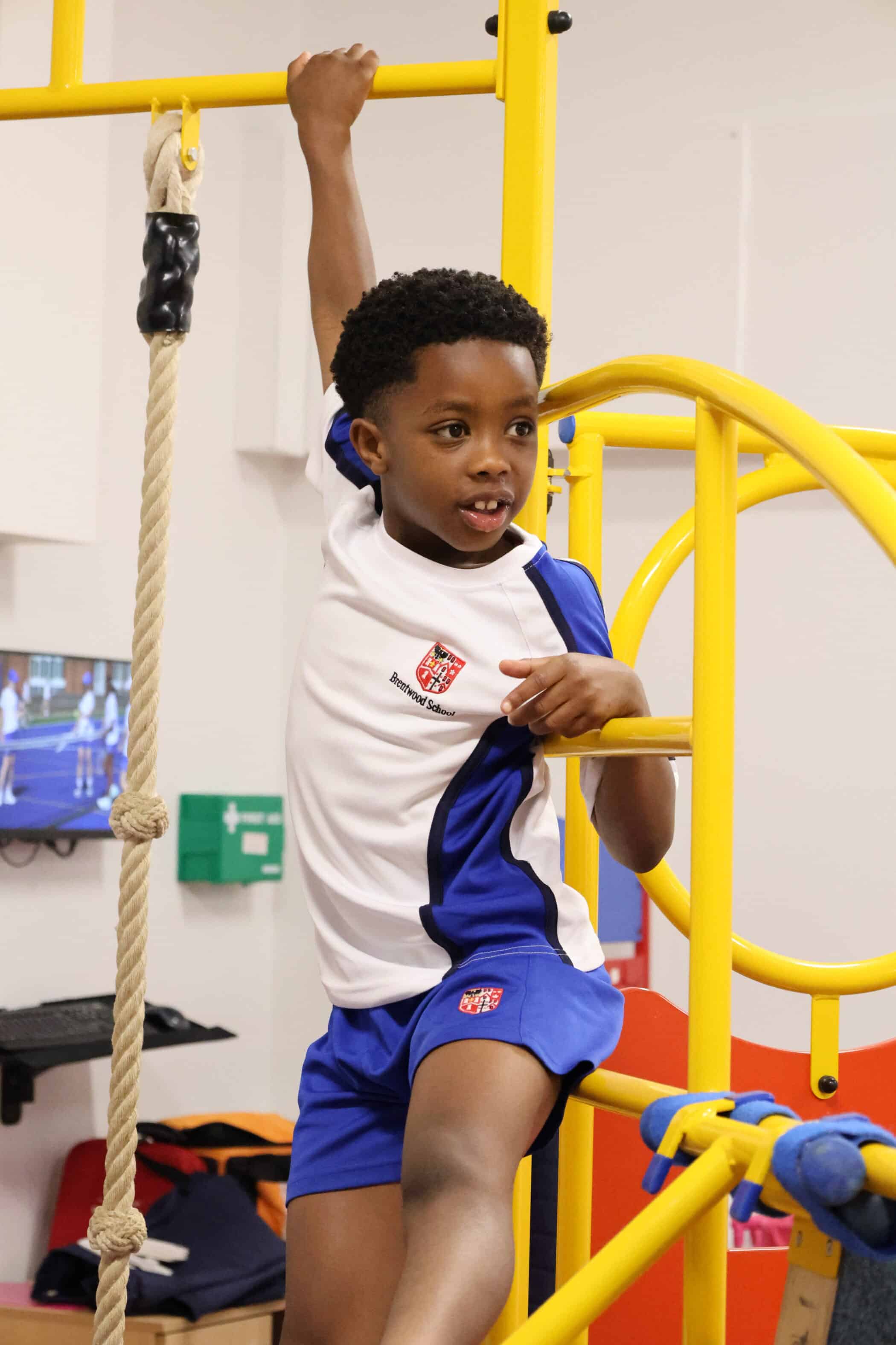 A young boy in a blue and white sports kit climbs yellow playground equipment indoors during Open Events, holding onto a bar with one hand and smiling.
