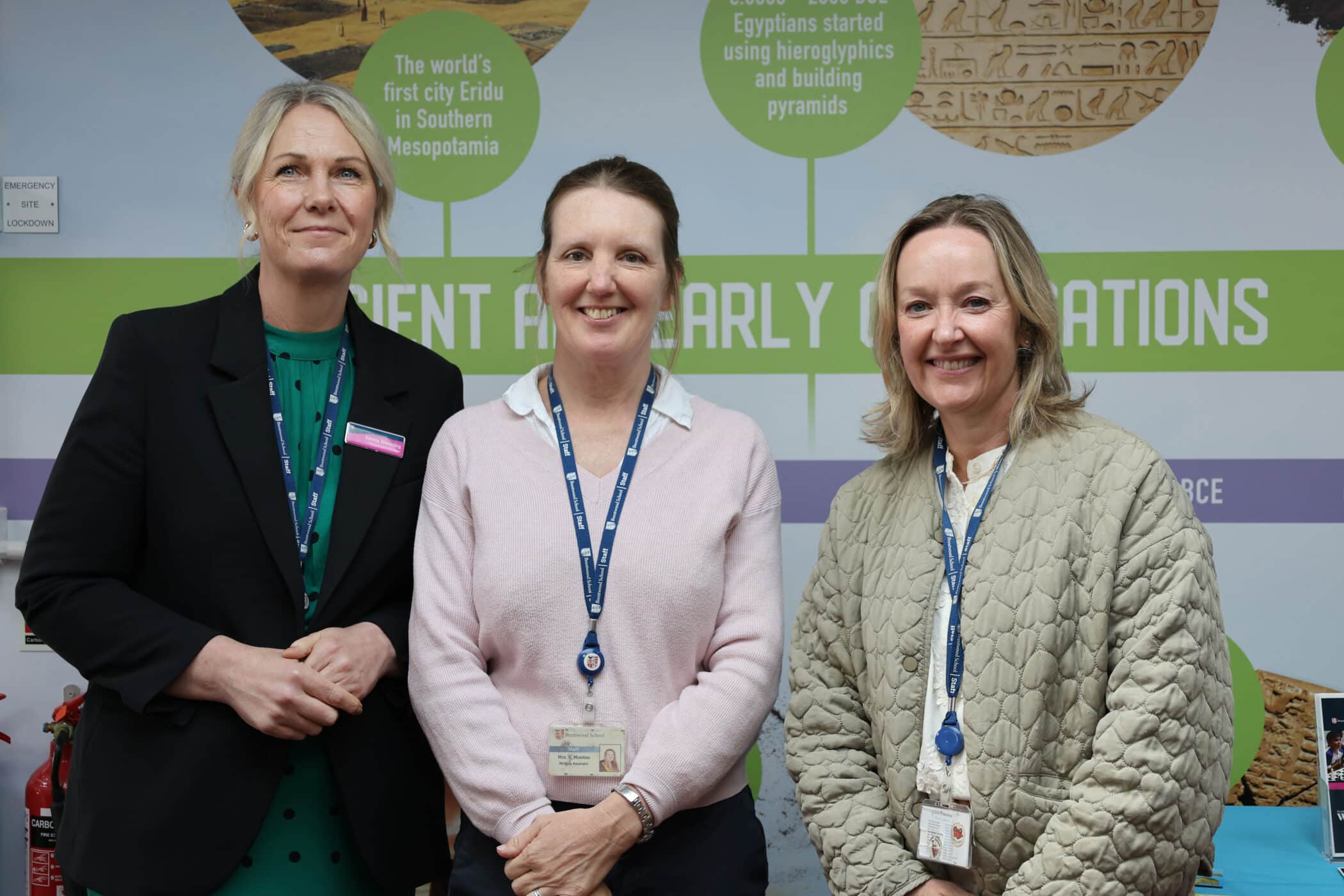 Three women wearing work badges and lanyards stand smiling in front of an educational display about ancient civilisations, including information on Mesopotamia and Egyptian pyramids, at one of the museum's exciting Open Events.