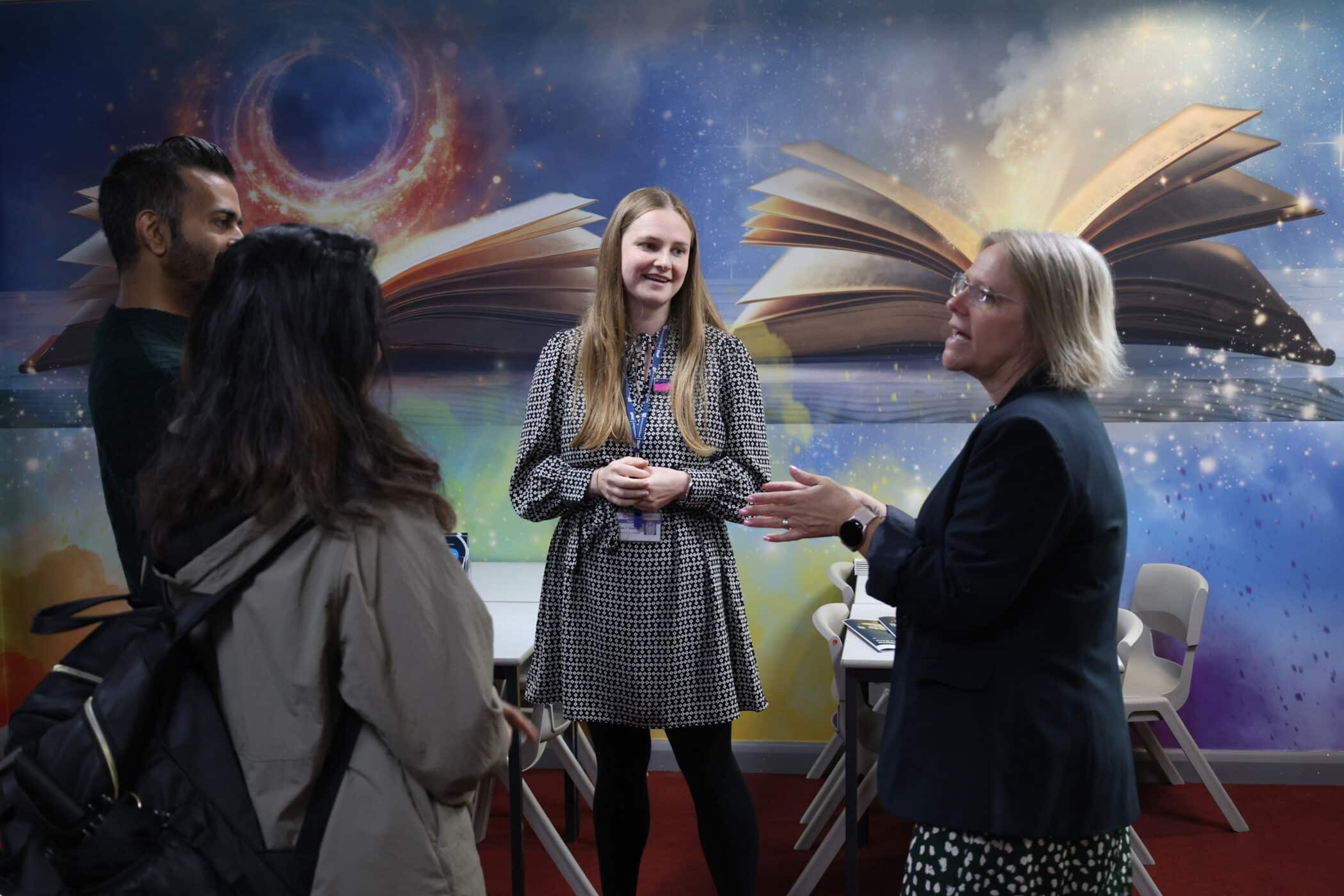 Four people stand and talk in a room with a colourful mural of open books and a cosmic background. During one of the Open Events, three face a smiling woman at the centre while another gestures as she speaks.