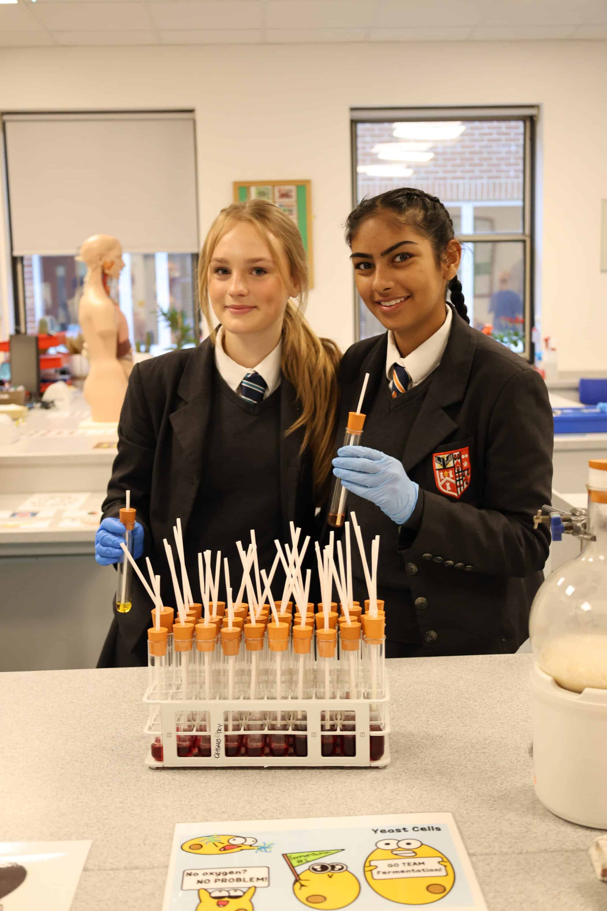 Two female students in school uniforms and blue gloves stand in a science laboratory at one of our Open Events, smiling whilst holding pipettes over a rack of test tubes filled with red liquid. Laboratory equipment and a mannequin are visible in the background.