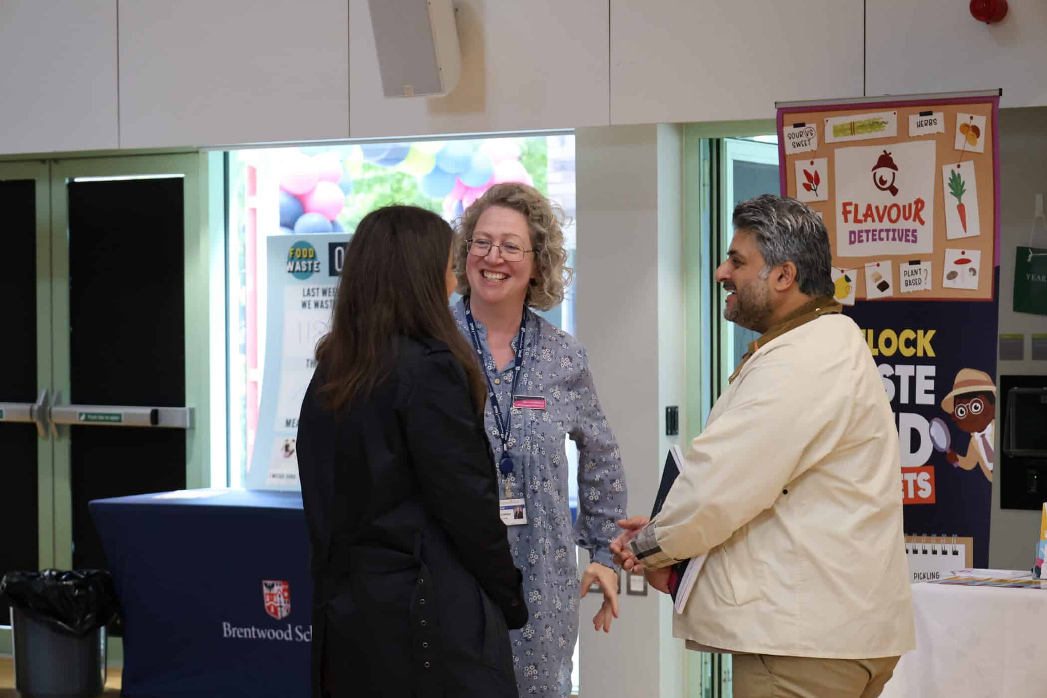 Three adults stand in a brightly lit room, smiling and talking together during one of Brentwood School’s Open Events. Behind them are colourful posters and banners, including Flavour Detectives and the school logo.