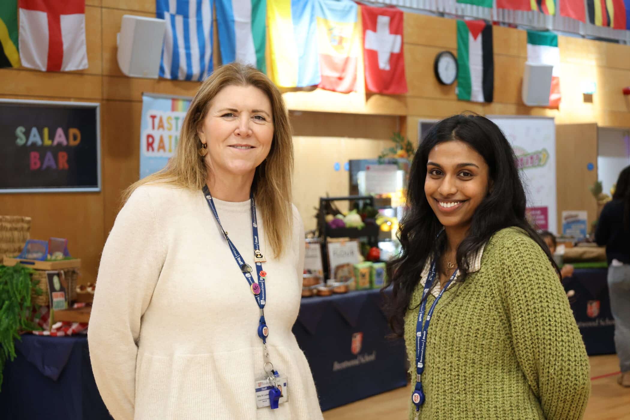 Two women, both wearing lanyards, stand smiling in front of food stalls and a wall decorated with international flags at an Open Event in a brightly lit indoor space.