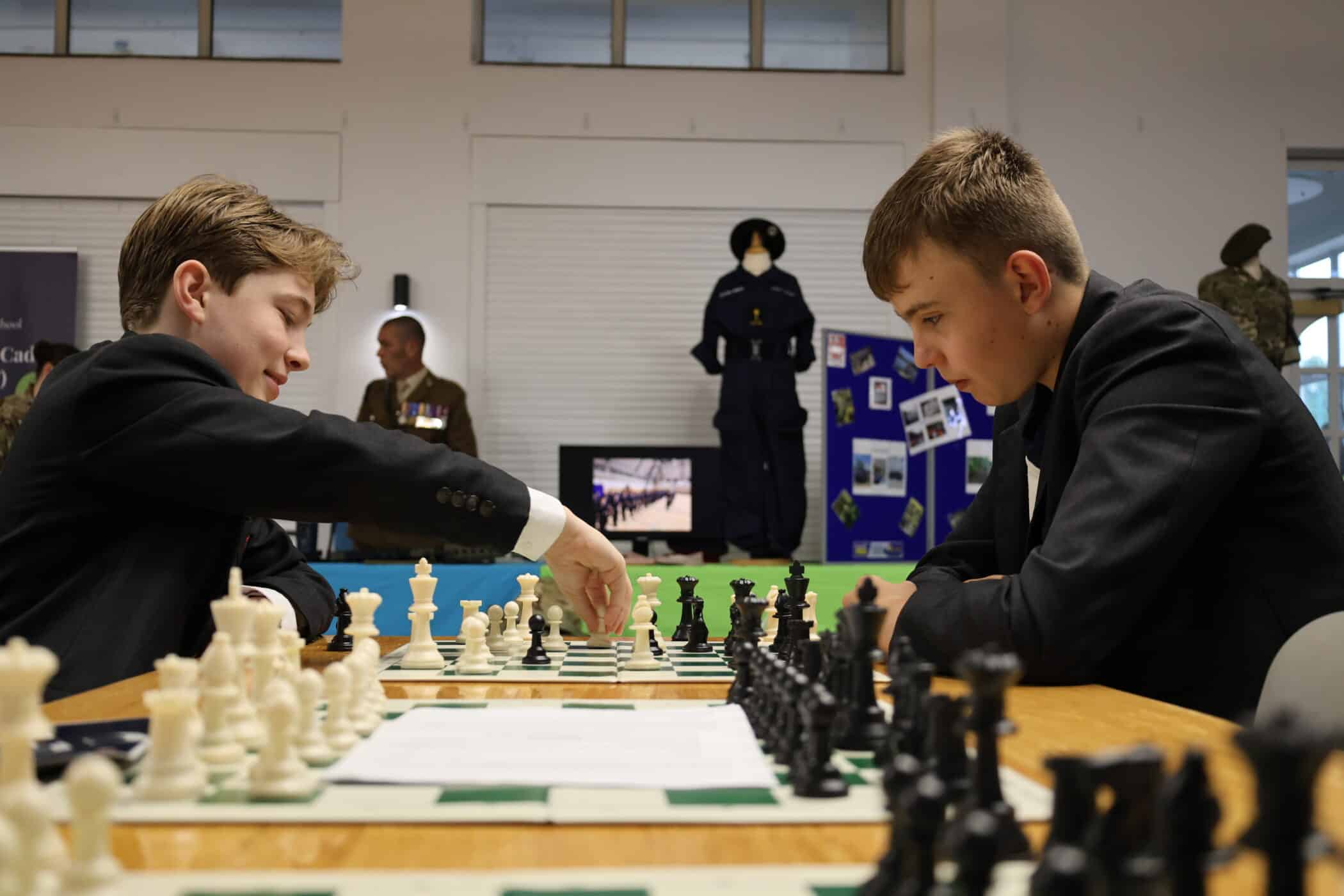 Two boys in school uniforms play chess at a table during Open Events. Behind them, mannequins in military uniforms and informational displays are visible in a brightly lit room.