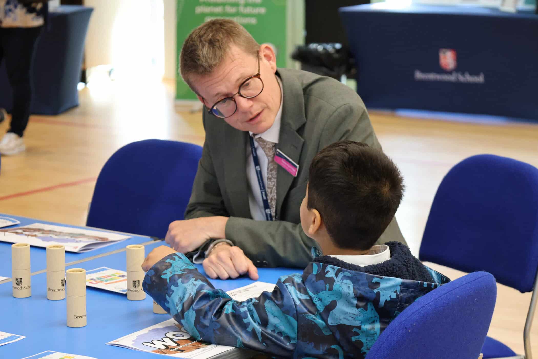 At an Open Events session, a man in a suit and glasses talks to a young boy in a blue jacket. They are seated at a table with papers, tubes, and colouring supplies in a brightly lit indoor setting.