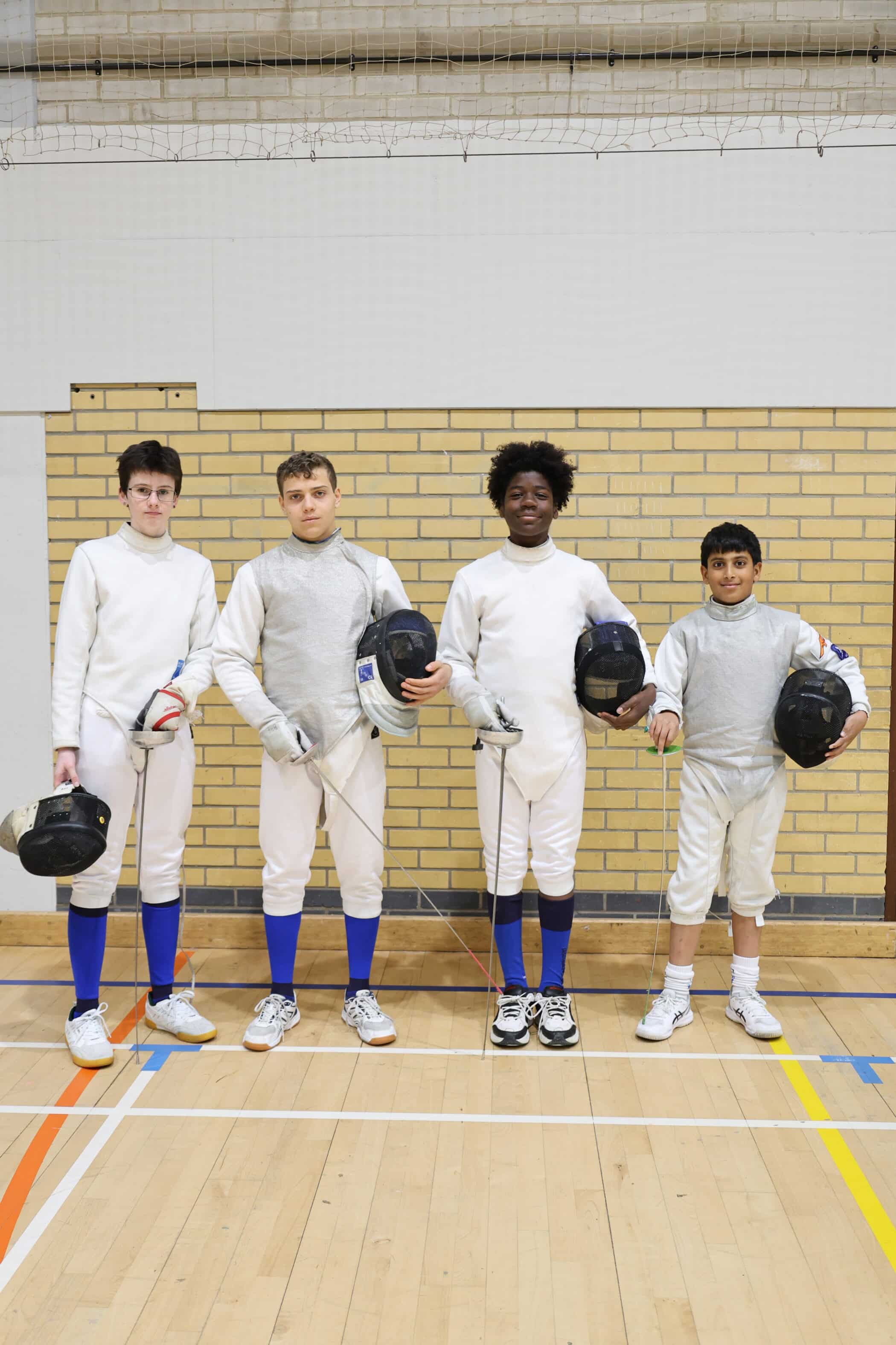 Four young fencers stand indoors on a gym floor, wearing white fencing kit and holding their masks. They face the camera side by side against a yellow brick wall, ready to compete in upcoming Open Events.