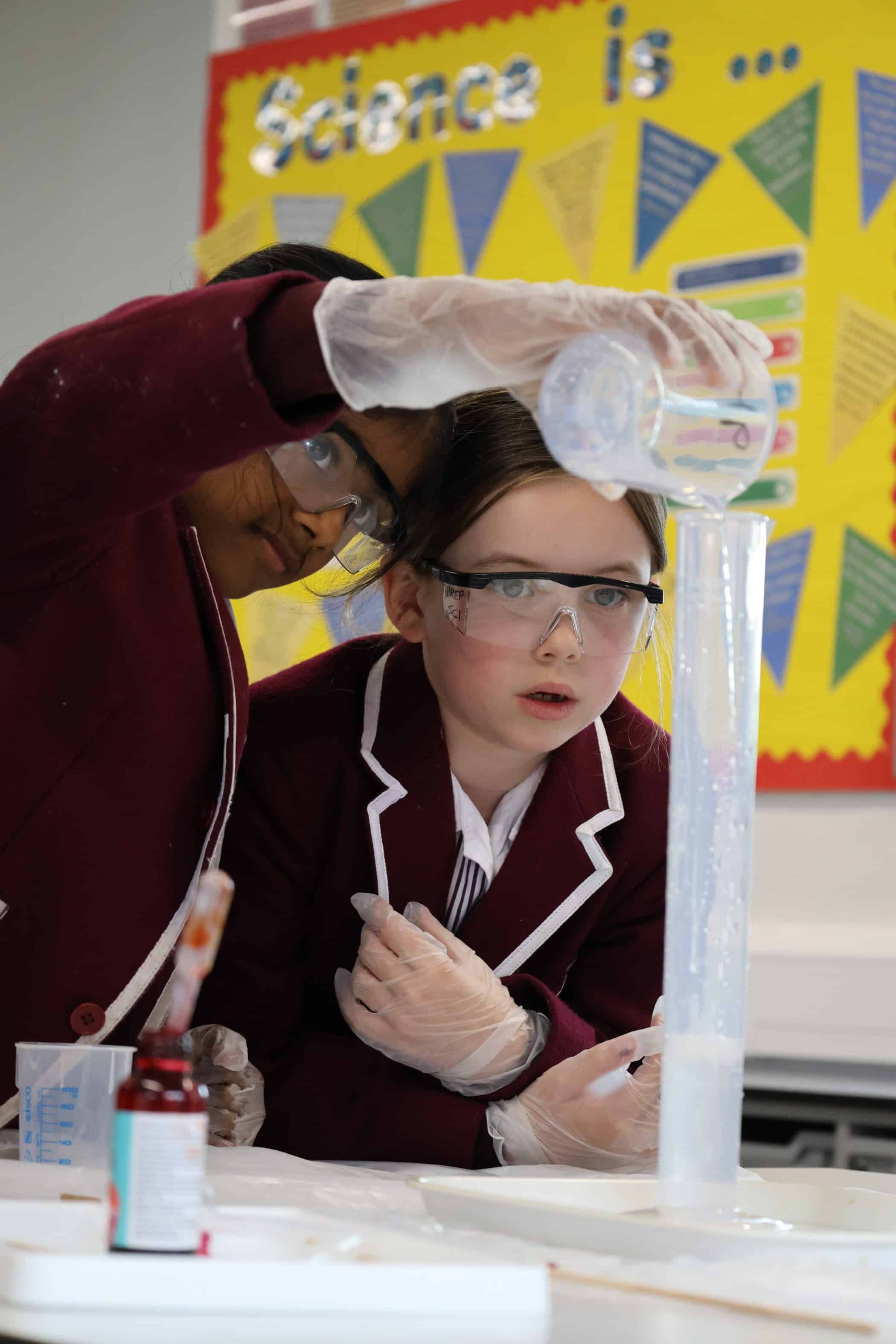 Two students wearing safety goggles and maroon blazers pour liquid into a tall graduated cylinder during a science experiment at one of our Open Events. A colourful noticeboard with Science is... is visible in the background.