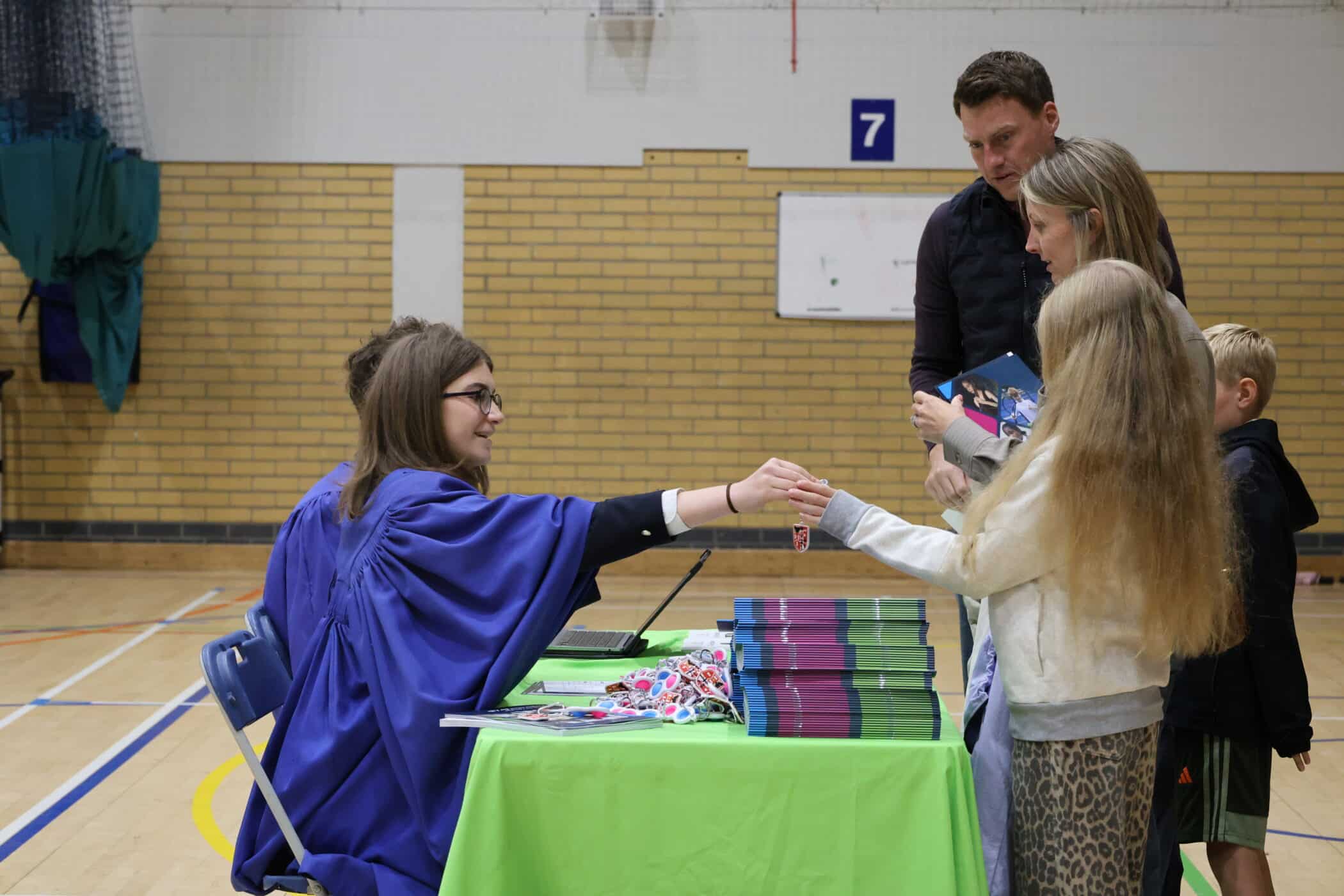 At an Open Events celebration in a gymnasium, a woman in a blue graduation gown sits at a table with stacked books, handing an item to a girl with long blonde hair whilst a man, woman, and young boy stand nearby.