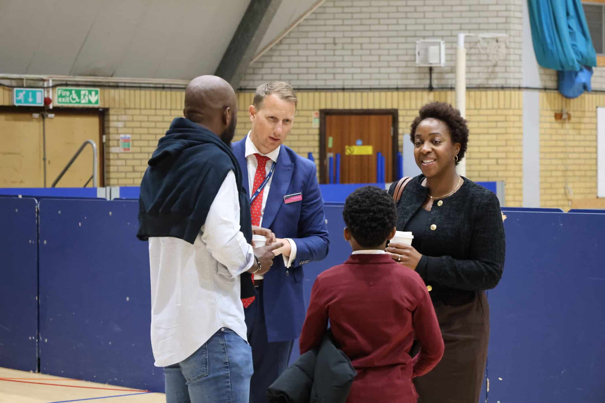 At an Open Events gathering, a man in a blue suit chats with a woman, another man, and a child in a maroon jumper inside a sports hall. The adults hold cups as they converse, with blue barriers and sports equipment visible in the background.