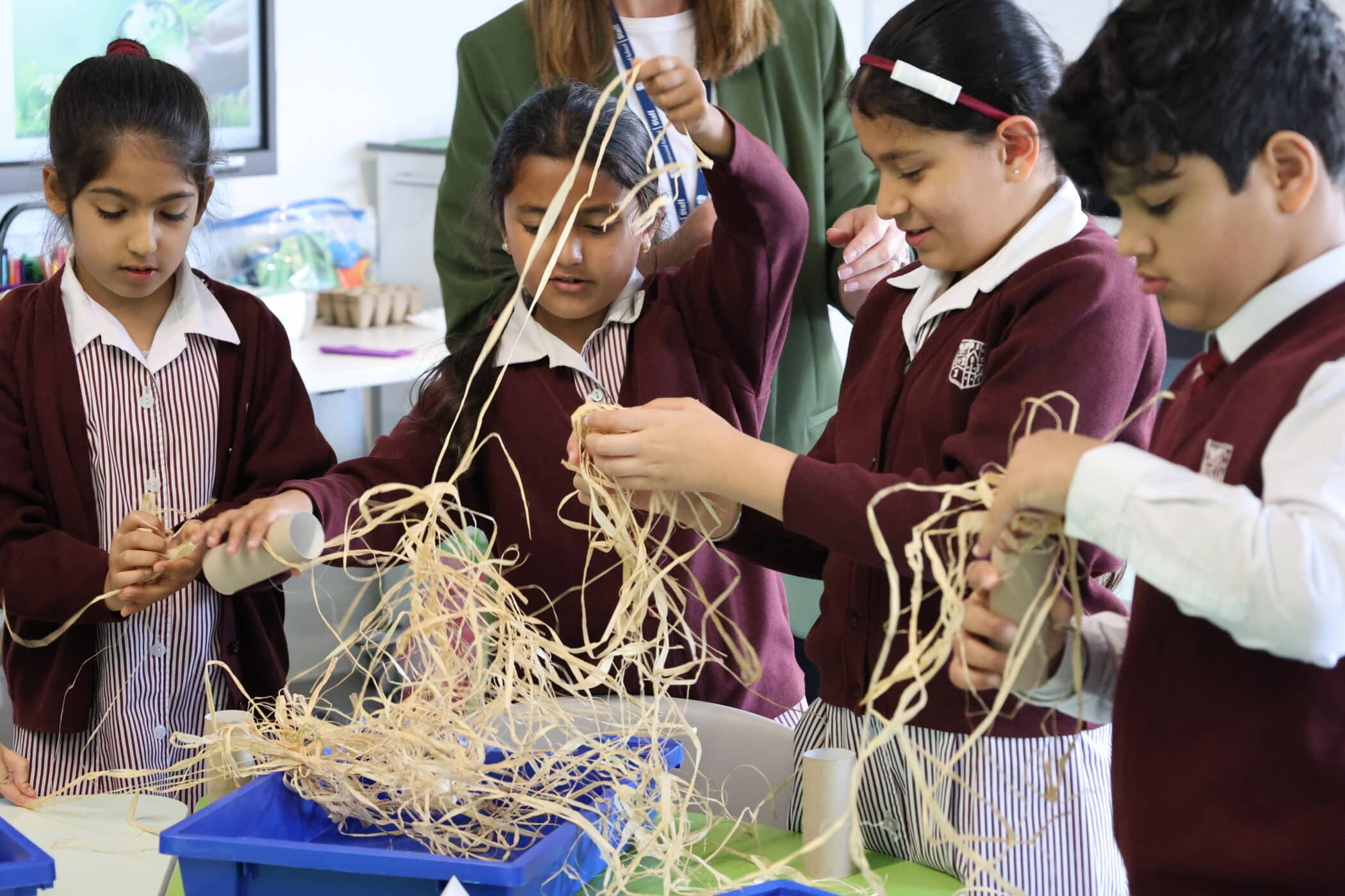 Four children in school uniforms work together with straw-like craft materials and cardboard tubes at a classroom table, as an adult oversees the activity in the background—capturing the hands-on creativity seen at our Open Events.