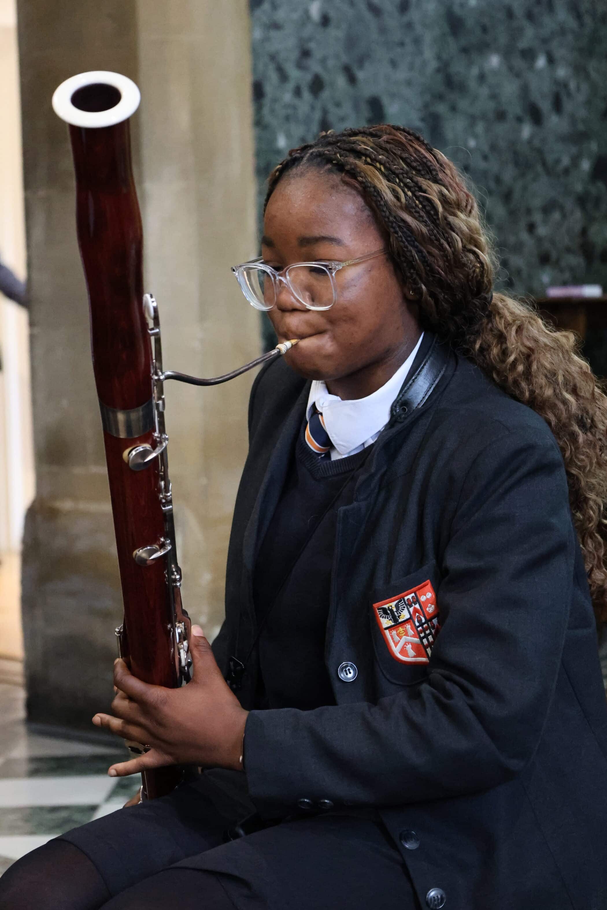A young musician in a school uniform and glasses plays a bassoon indoors, concentrating on her performance during one of the school's Open Events.