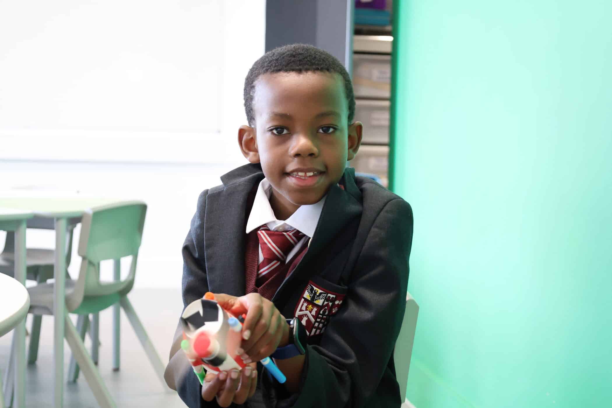 A young boy in a school uniform sits at a desk in a classroom, smiling and holding a colourful craft project—highlighting the creativity fostered during Open Events. The background features green walls and shelves with supplies.